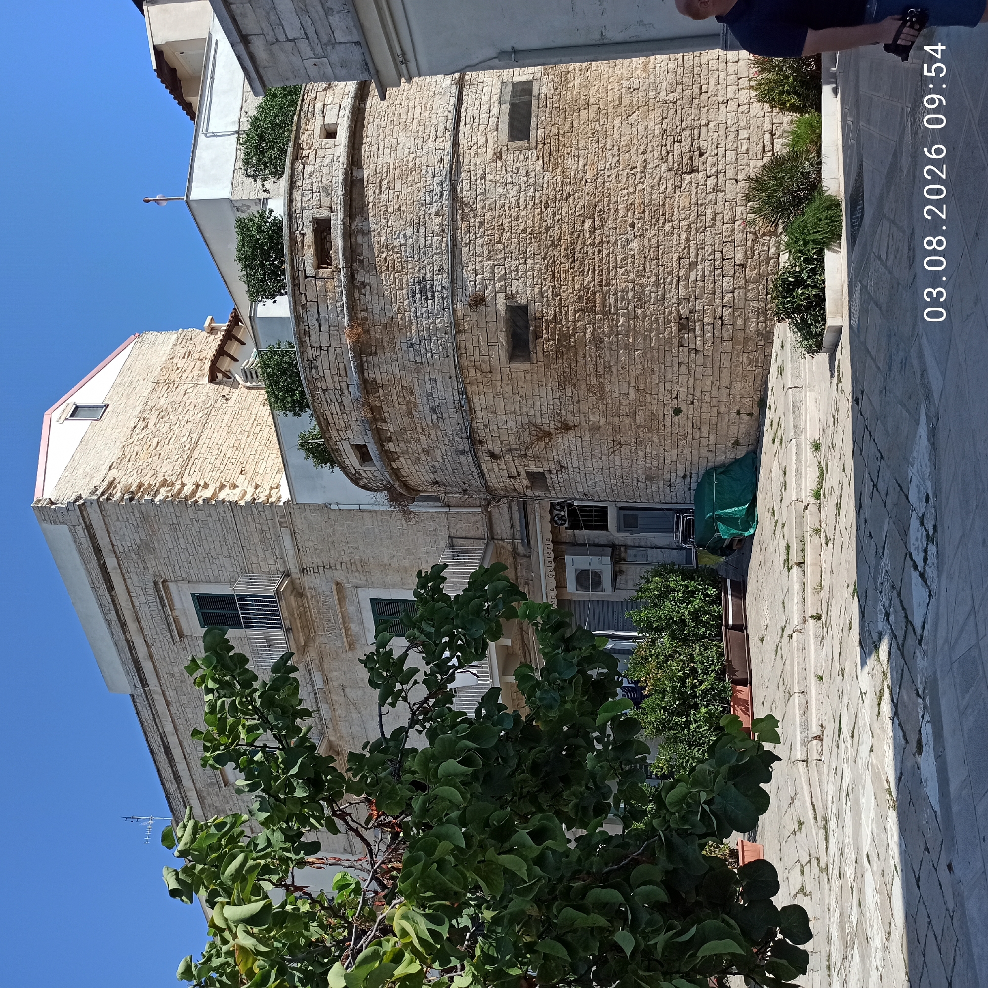 In an outdoor piazza in Ruvo di Puglia, Italy, a rounded stone structure stands prominently. A light-colored, multi-story building with balconies is visible behind and to the left of the stone structure. The piazza is paved with stone slabs and features several planters with green foliage. A tree with green leaves partially obscures the view of the buildings on the left. A person wearing a dark shirt and denim shorts is partially visible on the right side of the image. A timestamp "03.08.2026 09:54" is overlaid on the lower right portion of the image. The sky is clear and blue.