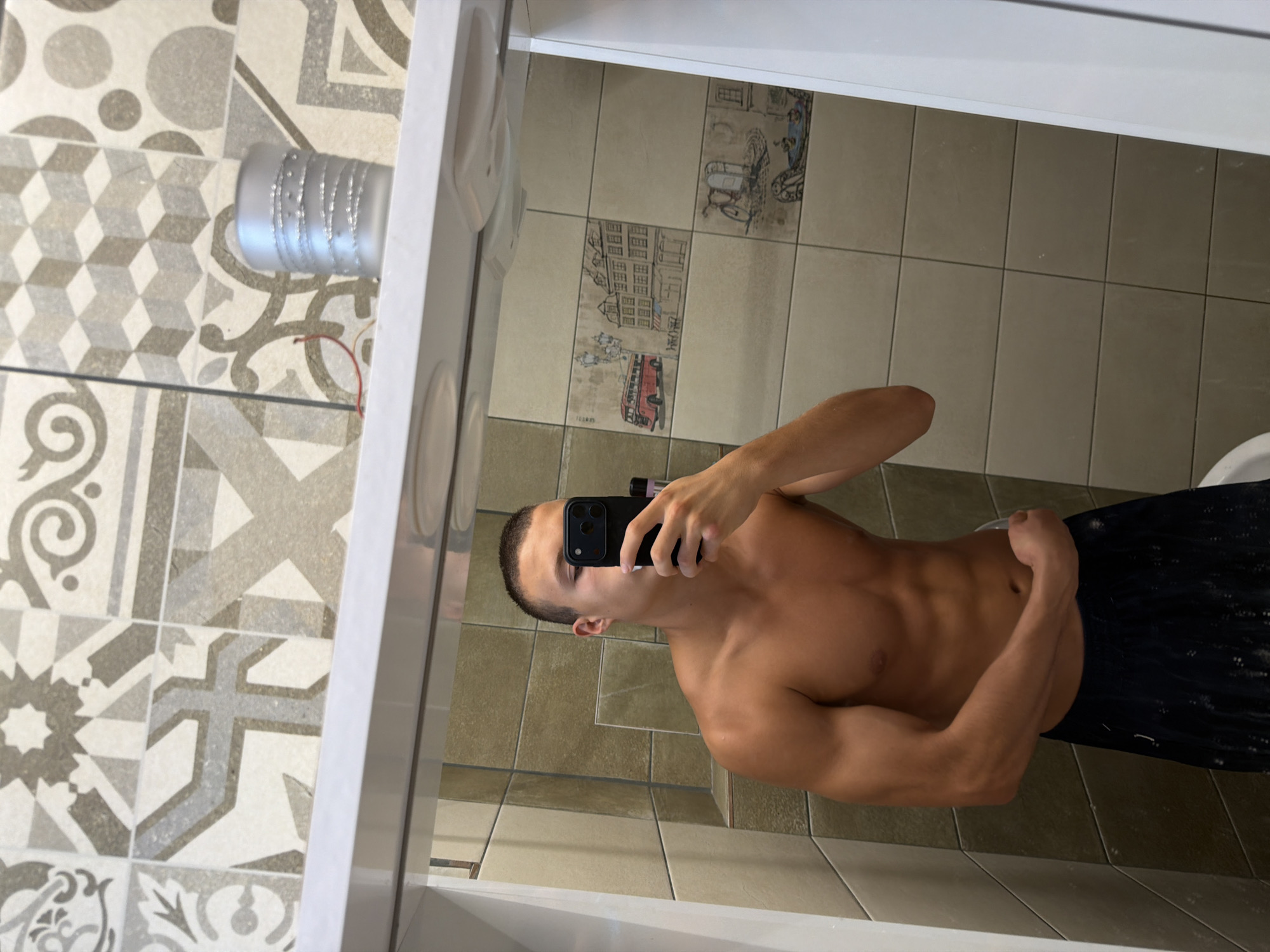 This is a mirror selfie taken by a young adult male in a bathroom. He is shirtless, wearing dark shorts, and holding a smartphone in his right hand to take the picture. The bathroom features tiled walls with a decorative pattern. The male appears to be in good physical condition, with visible musculature. The setting is domestic, and there is no indication of it being in Burgas, Bulgaria, or any specific city.