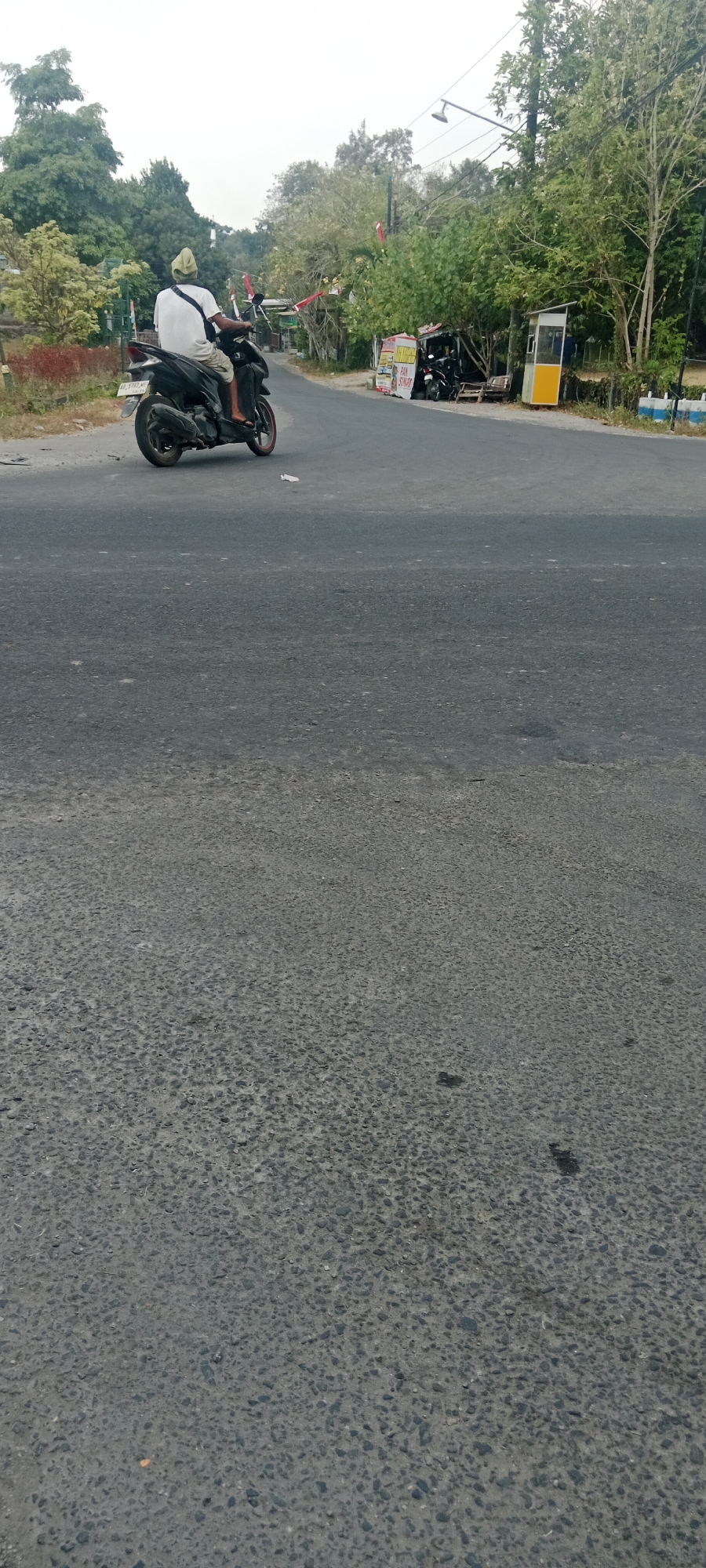 In Maguwoharjo, Indonesia, a person is riding a black scooter on a paved road. The rider, facing away from the camera, wears a white shirt, light-colored shorts, and a light-colored head covering. The scooter has a license plate visible on the rear. To the right of the road, a small, open-air stall with a yellow and white structure is present, next to a few parked motorcycles. Trees and greenery line the side of the road in the background. The sky is overcast.