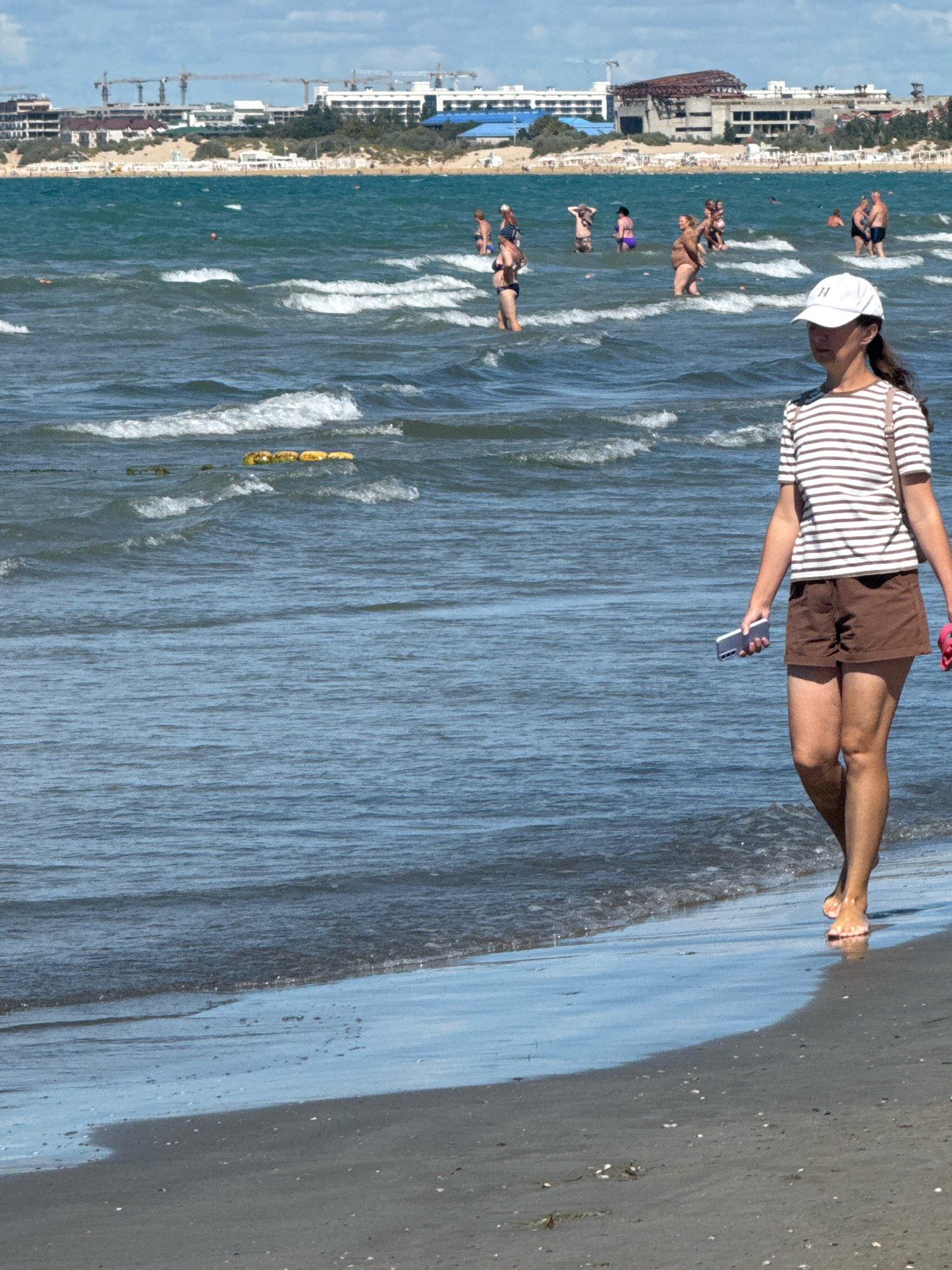 A woman walks on the beach in Anapa, Russia, near the Black Sea. The woman is wearing a striped shirt, brown shorts, and a white cap. She is holding a smartphone in her left hand. In the background, several people are wading in the water. The shoreline is sandy, and there are some waves. Buildings and construction cranes are visible in the distance along the coast.