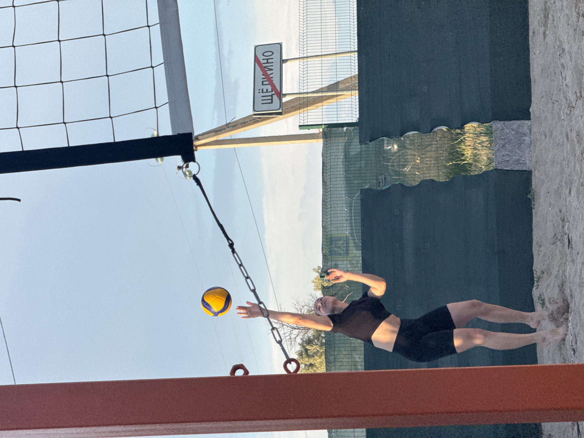 A young woman is in mid-air, jumping to hit a volleyball on a beach volleyball court. She is wearing a black sports bra and black shorts, and her arms are extended upwards. The volleyball is yellow and blue and is above her right hand. The court is made of sand and is enclosed by a dark green fence. A red support post is visible on the left side of the image, and a volleyball net is stretched across the top of the frame. In the background, a sign with Russian text reading "ЩЁЛКИНО" (Shchyolkino) is mounted on a wooden pole, with a red line through it, indicating a boundary or entrance into the town. The sky is a clear light blue, suggesting it is either early morning or late afternoon.