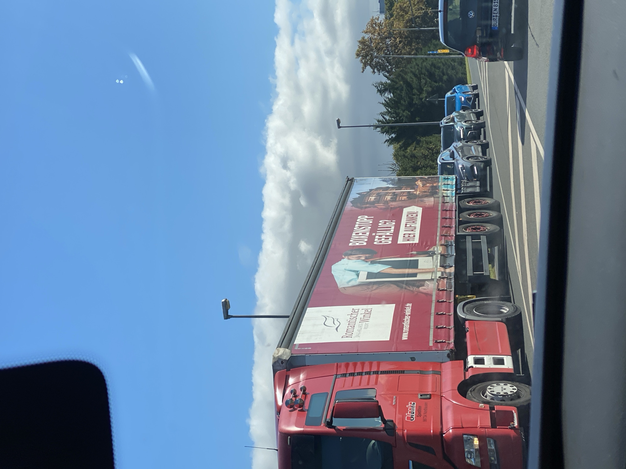 The image depicts a red semi-truck with a trailer driving on a road in Nordhausen, Germany, on a sunny day with some clouds. The trailer is adorned with a large advertisement for "Romantischer Winkel," a spa and wellness resort, with the website "www.romantischer-winkel.de" prominently displayed. The truck has "Blanke Spedition 99734 Nordhausen" printed on the cab. Several cars are visible behind the truck, also driving on the road. The scene appears to be a typical daytime road view in a populated area, likely a highway or a main thoroughfare.