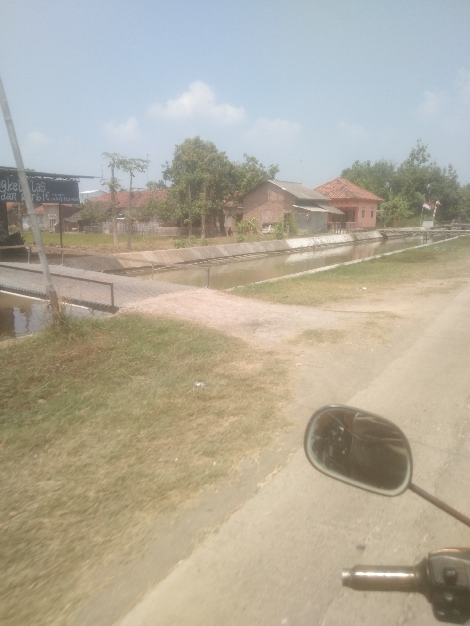 The image shows a canal with a concrete embankment and a small bridge crossing it. On the left side of the canal, there is a sign that reads "Ngket Las dan Arsit" followed by a phone number. Beyond the canal, there are several houses and trees. The sky is clear with a few clouds. The foreground shows a dirt road and a motorcycle's rearview mirror and handlebars, suggesting the photo was taken from a moving vehicle. The scene is indicative of a rural or semi-rural area in Desa Sukra, Indonesia, likely during daytime.