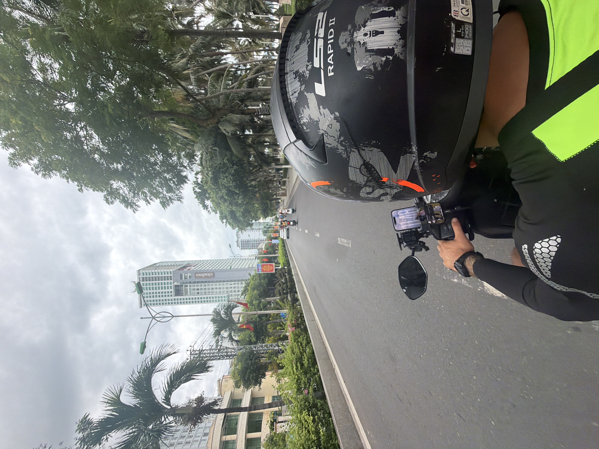 The image captures a first-person perspective from a motorcycle on a street in Nha Trang, Vietnam. The rider is wearing a black LS2 Rapid II helmet with a distressed graphic design and orange accents. A smartphone is mounted on the handlebars, displaying a time of "11:48". The rider's left arm, with a black watch, is visible.
The street is paved and appears to be a multi-lane road with a concrete median barrier lined with greenery. To the left, there are buildings, including a light-colored building with balconies and a taller, modern skyscraper with a green facade and "Citadines" branding. Palm trees and other lush vegetation line the street, creating a tropical atmosphere. In the distance, other motorcycles and a white car are visible on the road. The sky is overcast with grey clouds. The scene is consistent with the coastal urban environment of Nha Trang.