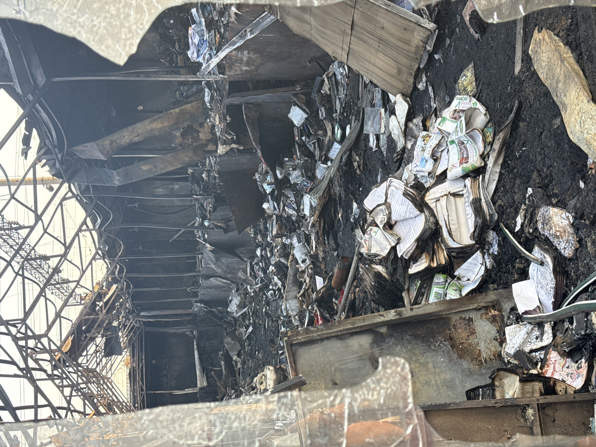 The image shows the interior of a building, likely a commercial space or warehouse, in Kyiv, Ukraine, that has been heavily damaged by fire. The roof structure is exposed and appears warped and blackened, with exposed metal beams. The walls are charred, and debris, including burnt paper, plastic, and assorted objects, covers the ground. Several stacks of books and papers are visible, some appearing to be somewhat intact but water-damaged and soiled. The overall scene suggests destruction and disarray. The foreground is partially obscured by what appears to be cracked glass, possibly from a window or door, through which the photograph was taken.