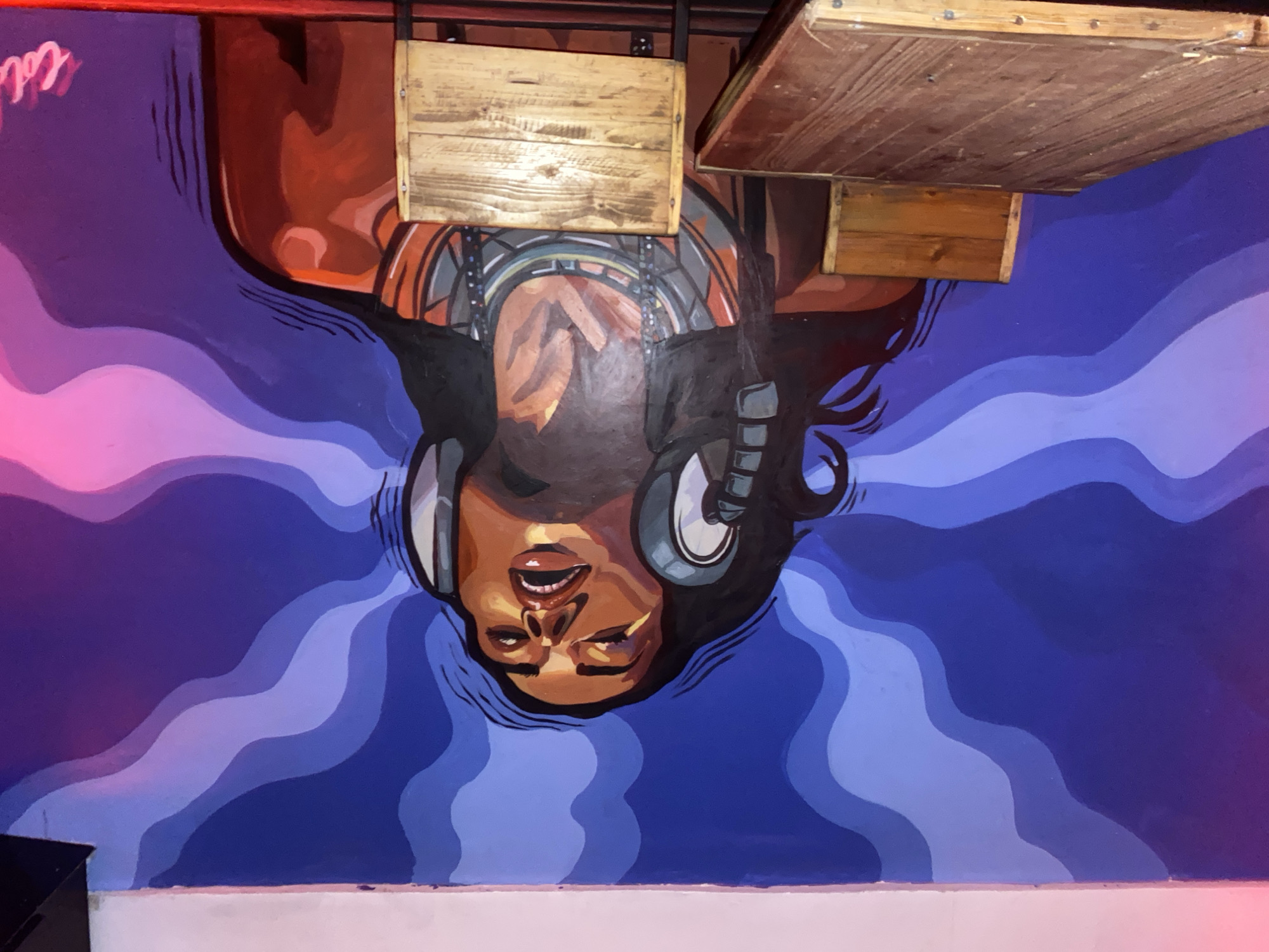 The image shows a mural on a wall in Howayi, Nigeria. The mural depicts a woman wearing headphones, with her eyes closed and mouth open, as if singing or listening to music. She has dark hair and is wearing a large necklace. The background of the mural is a gradient of blue and purple waves, resembling sound waves or smoke. In the foreground, there are two wooden chairs and a wooden table, suggesting this is a seating area within a venue such as a bar or restaurant. The lighting in the scene appears to be from colored lights, with pink hues visible on the right side of the mural. A signature, "Coli," is visible on the lower right corner of the mural.
