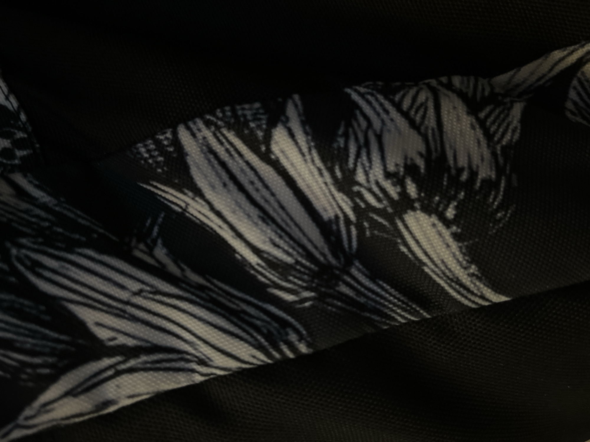 The image is a close-up, abstract shot of a black fabric with a white and black palm leaf pattern. The fabric appears to be part of a bag or backpack, with a textured black material visible in the background. The lighting is dim, creating shadows and highlighting the texture of the fabrics. There are no people or animals visible in the image. The location in Kalynivka, Ukraine is not discernible from the visual information provided.