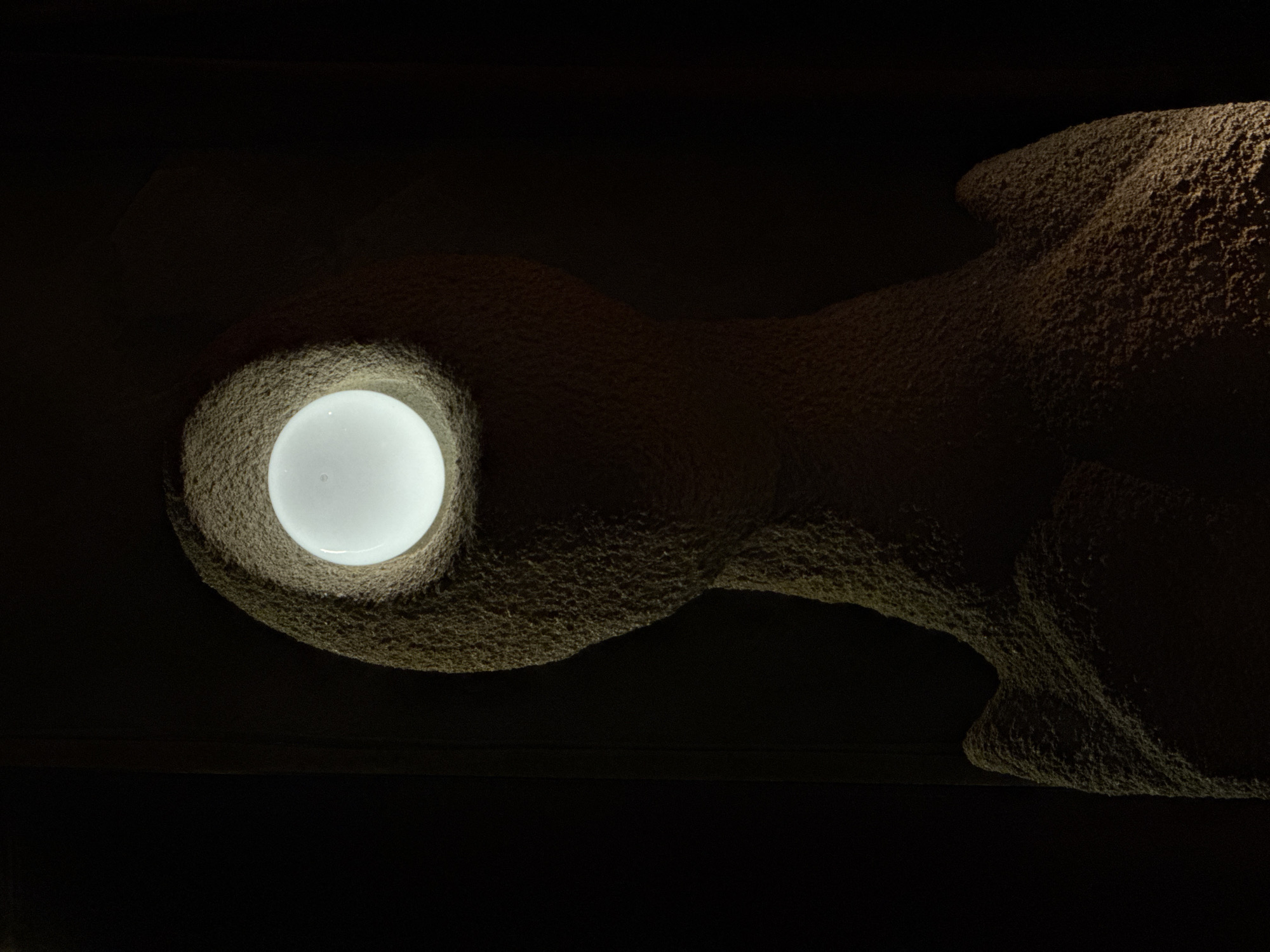 The image depicts a bulbous, textured, possibly stone or concrete structure with a rounded recess containing a glowing, spherical light. The structure is positioned against a dark background, with the light illuminating the interior of the recess and casting a subtle glow on the surrounding surface. The texture of the structure appears coarse and granular. The overall scene is dimly lit, relying primarily on the light source within the recess. I am unable to determine the location in Moscow, Russia.