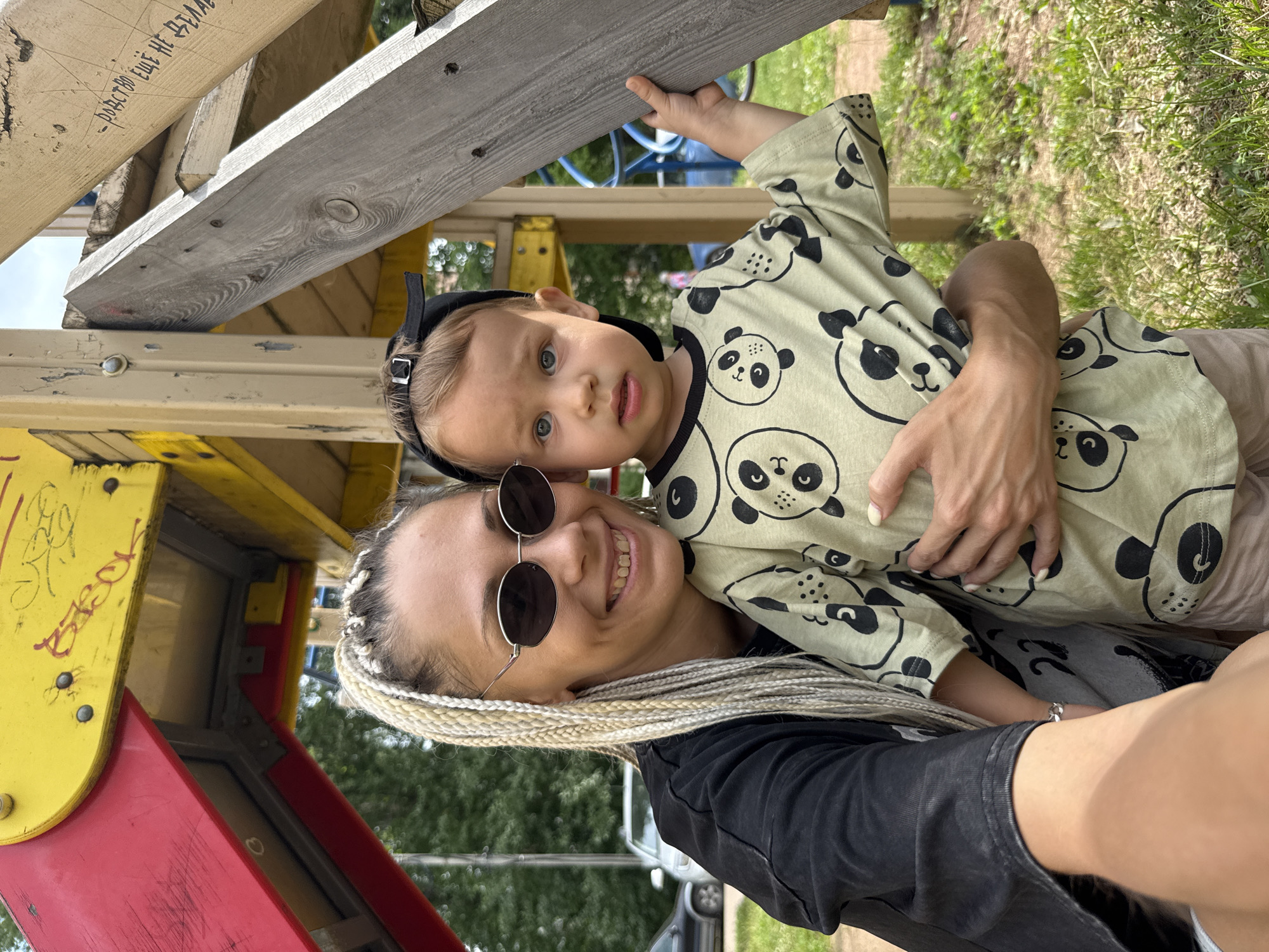 A woman with blonde braided hair and sunglasses smiles while holding a young child in her arms. The child, wearing a light green shirt with a panda print and a black baseball cap, looks towards the camera. They are standing outdoors near a yellow and red playground structure made of wood and metal. The environment includes grass and trees, with some cars visible in the background, suggesting a park or residential area. The image appears to be a candid shot taken on a sunny day. There is some graffiti on the playground equipment.
