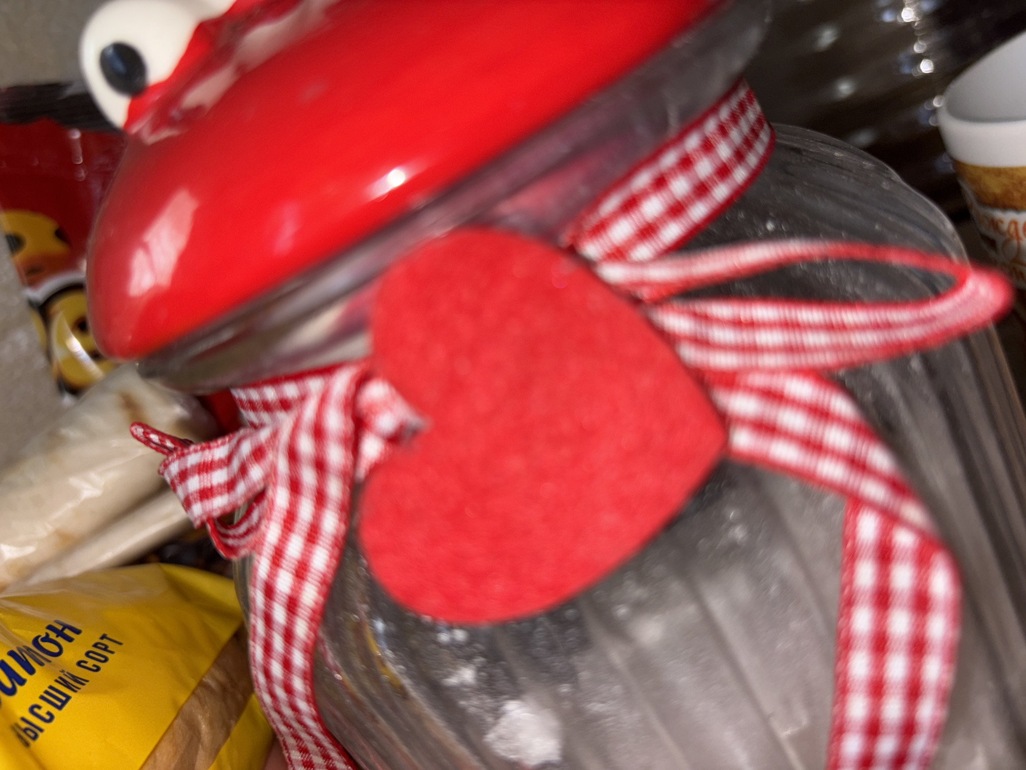 The image shows a close-up view of a red, novelty jar lid with eyes, possibly shaped like a character. A red and white gingham ribbon is tied around the neck of the jar, with a small, red, heart-shaped embellishment attached to the ribbon. The jar itself appears to be made of glass and is filled with something that is not clearly visible due to the focus and clarity of the image. To the left of the jar, a yellow plastic bag with some text on it is partially visible. The text is in blue Cyrillic script and appears to say "Умный сорт" or similar. The overall scene is a still life arrangement, likely indoors, with the focus on the decorative jar and its accessories. The lighting is soft, and there are some out-of-focus highlights in the background, suggesting a home or craft environment. No people are visible in the image.