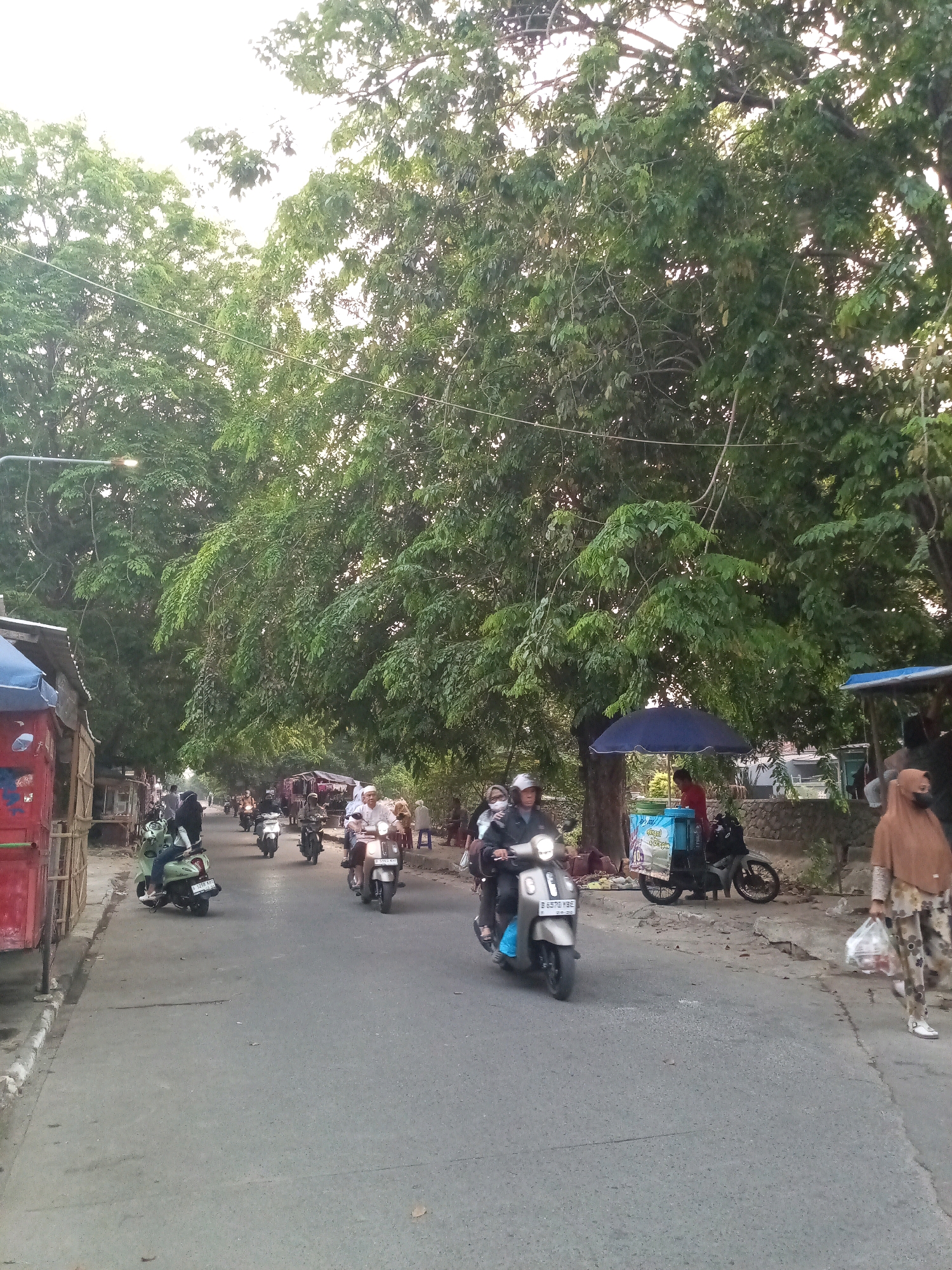 The image depicts a street scene in the city of Bekasi, Indonesia, characterized by a moderately wide asphalt road lined with lush trees that create a shaded canopy. Several motorcycles are present, with riders navigating the road. On the left side of the road, a red and blue temporary stall is visible, with wooden supports. Further down the road, on the right side, a blue umbrella shades a food cart or vendor.
People are observed riding motorcycles, with varying degrees of clothing that suggest a warm climate. One person in the foreground on the right is walking, carrying a plastic bag. The overall atmosphere is that of a bustling local street, likely in a residential or commercial area of Bekasi. The time of day appears to be late afternoon or early evening, as indicated by the quality of light filtering through the trees.