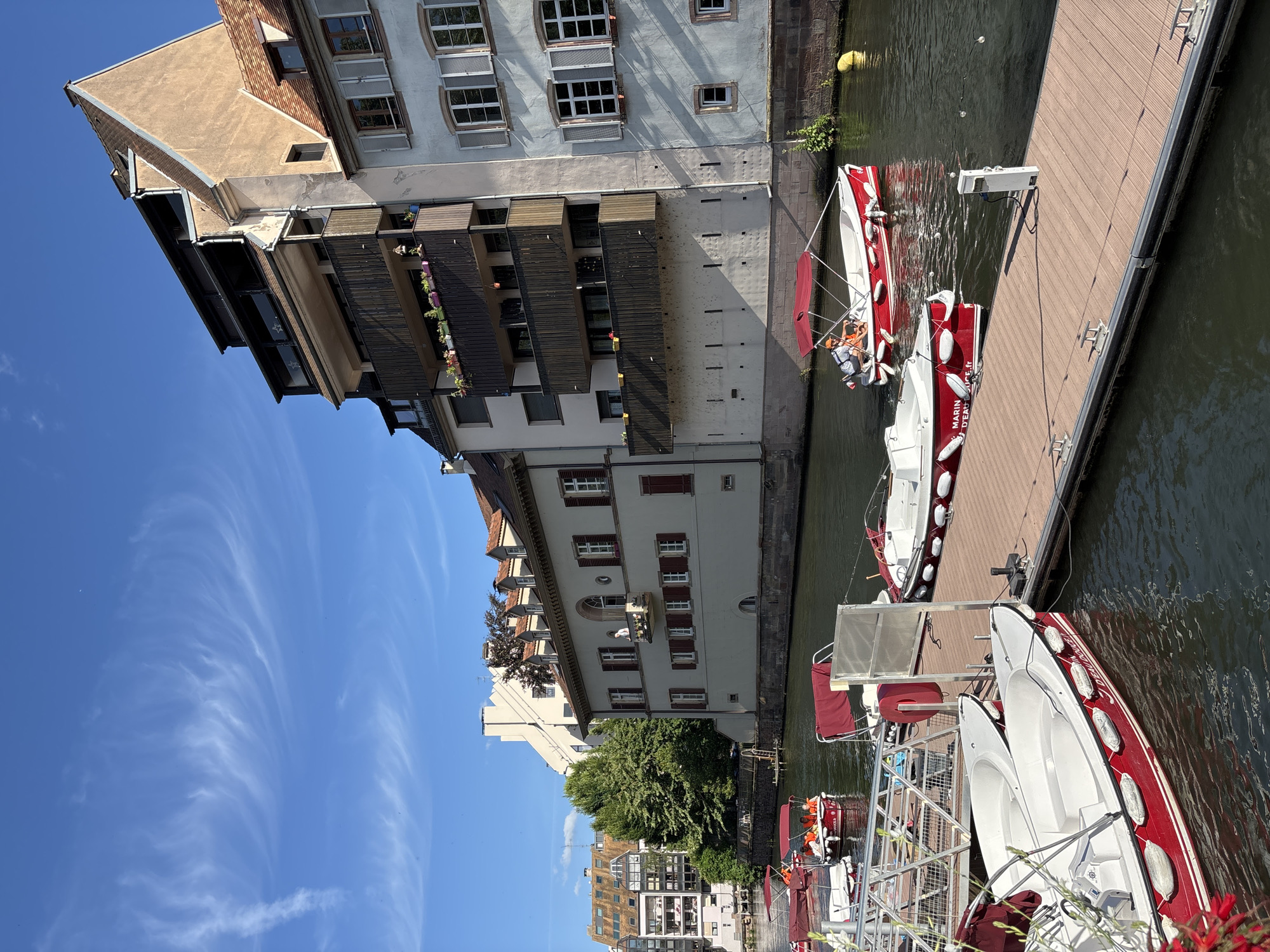 This image captures a sunny day along a canal in Strasbourg, France. The scene is dominated by a wooden pier where several small, red and white motorboats are docked. The boats are designed for recreational use, with some featuring red canopies. One boat in the foreground has the words "MARIN D'EAU DOUCE" and ".fr" visible on its hull. Another boat further out on the canal has two individuals on board, one appearing to be steering.
The canal is lined with historic buildings. The architecture is varied, with some structures featuring traditional Alsatian tiled roofs and multiple stories with balconies. One building has a light-colored facade with multiple windows and red shutters. Another building, closer to the left side of the frame, appears to be more modern with balconies and several floors. Trees are visible between the buildings, adding a touch of green to the urban landscape.
The sky is a clear, bright blue with wispy white clouds. The water of the canal is dark and reflects the buildings and sky. The overall impression is one of a peaceful and pleasant day in the city.