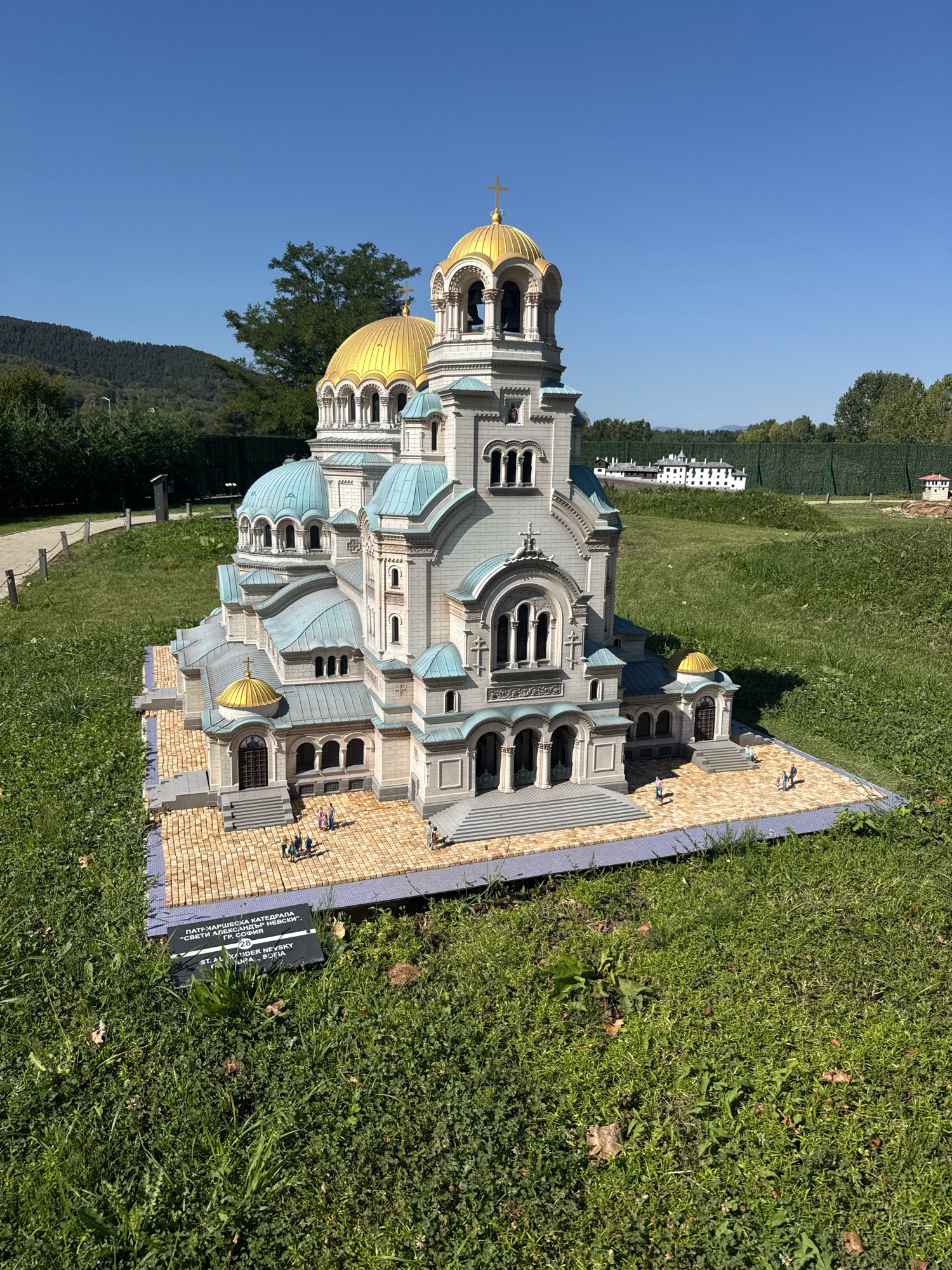 The image shows a detailed miniature model of the St. Alexander Nevsky Cathedral, a famous landmark in Sofia, Bulgaria. The model is set in an outdoor park-like environment with green grass surrounding it. The cathedral model is constructed with meticulous detail, featuring golden domes and light-colored walls with turquoise accents on the roofs. Small figures, representing people, are placed around the base of the model, giving a sense of scale. A sign is visible in front of the model, displaying text in Cyrillic and Latin alphabets, identifying the structure as "ST. ALEX. NEVSKY CATHEDRAL, SOFIA". In the background, there are trees and a distant view of buildings and hills, suggesting a sunny day with a clear blue sky. The location is identified as the city of Pravets, Bulgaria, implying this miniature park is situated there.