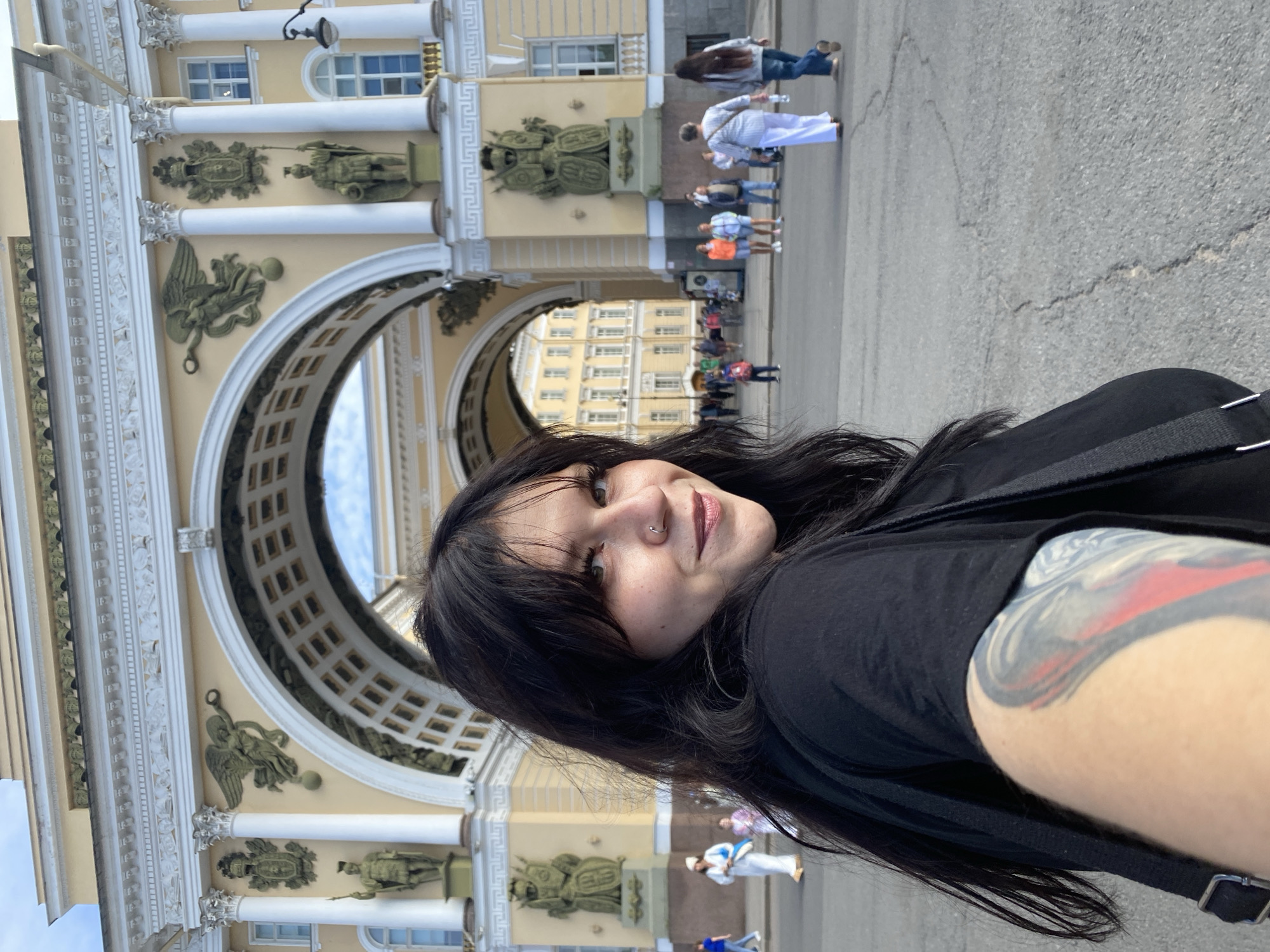 In the foreground, a young woman with long, dark hair and a nose ring poses for a selfie. She is wearing a black t-shirt and a black bag is slung over her shoulder, revealing a large, colorful tattoo on her arm. The woman is smiling, looking towards the camera. In the background, the iconic Arch of the General Staff Building in Palace Square, Saint Petersburg, Russia, is visible. The arch is adorned with sculptures and classical architectural details. Several people are walking in the square, some in the distance, blurred by the focus on the foreground subject. The ground is paved, and the sky is a light blue.