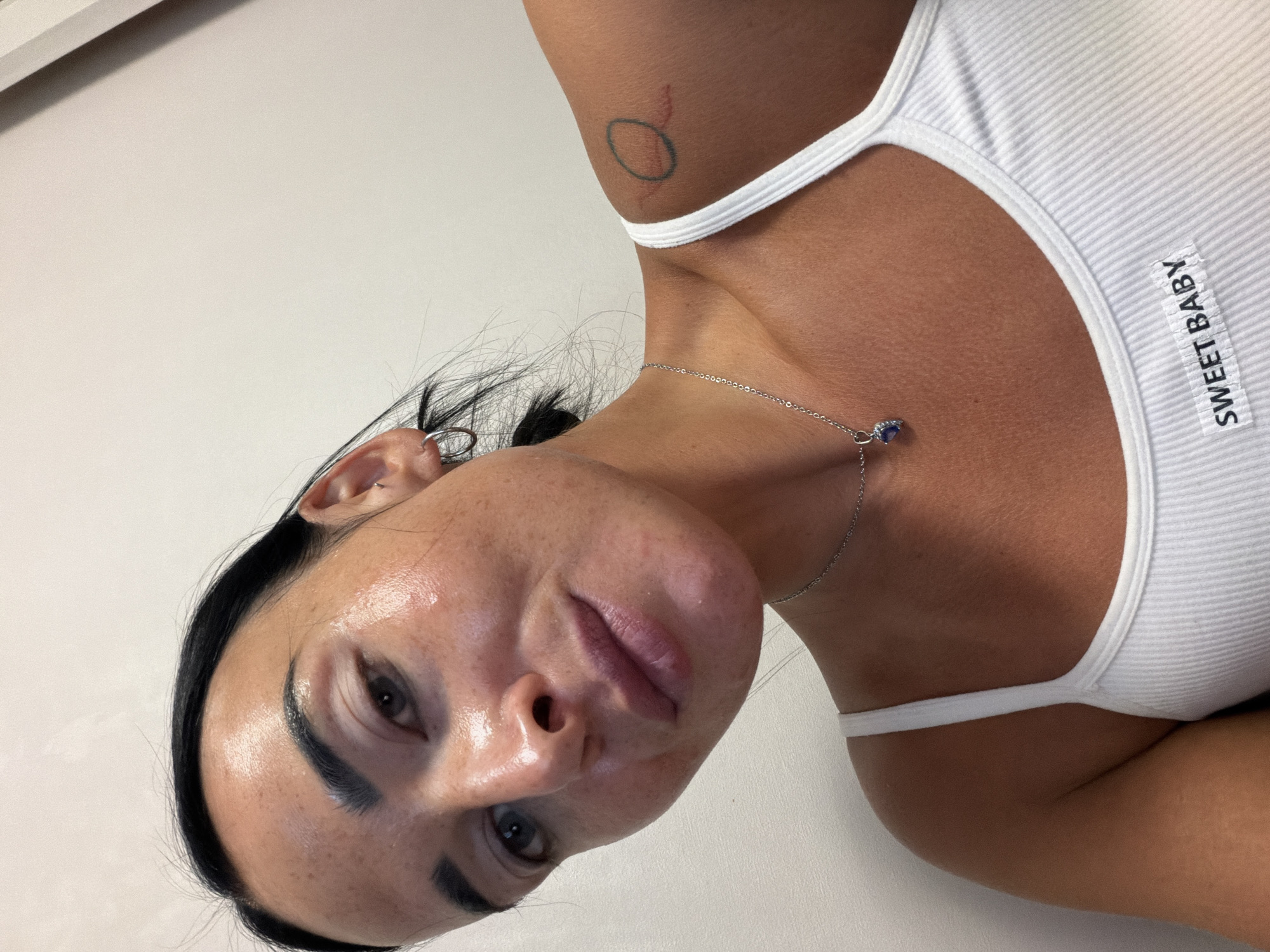 A woman with dark hair is shown in a close-up selfie. She is wearing a white ribbed tank top with "SWEET BABY" printed on the front. A silver necklace with a blue pendant is around her neck. She has a tattoo on her shoulder. The background is a plain white wall. The location is not discernible from the image.