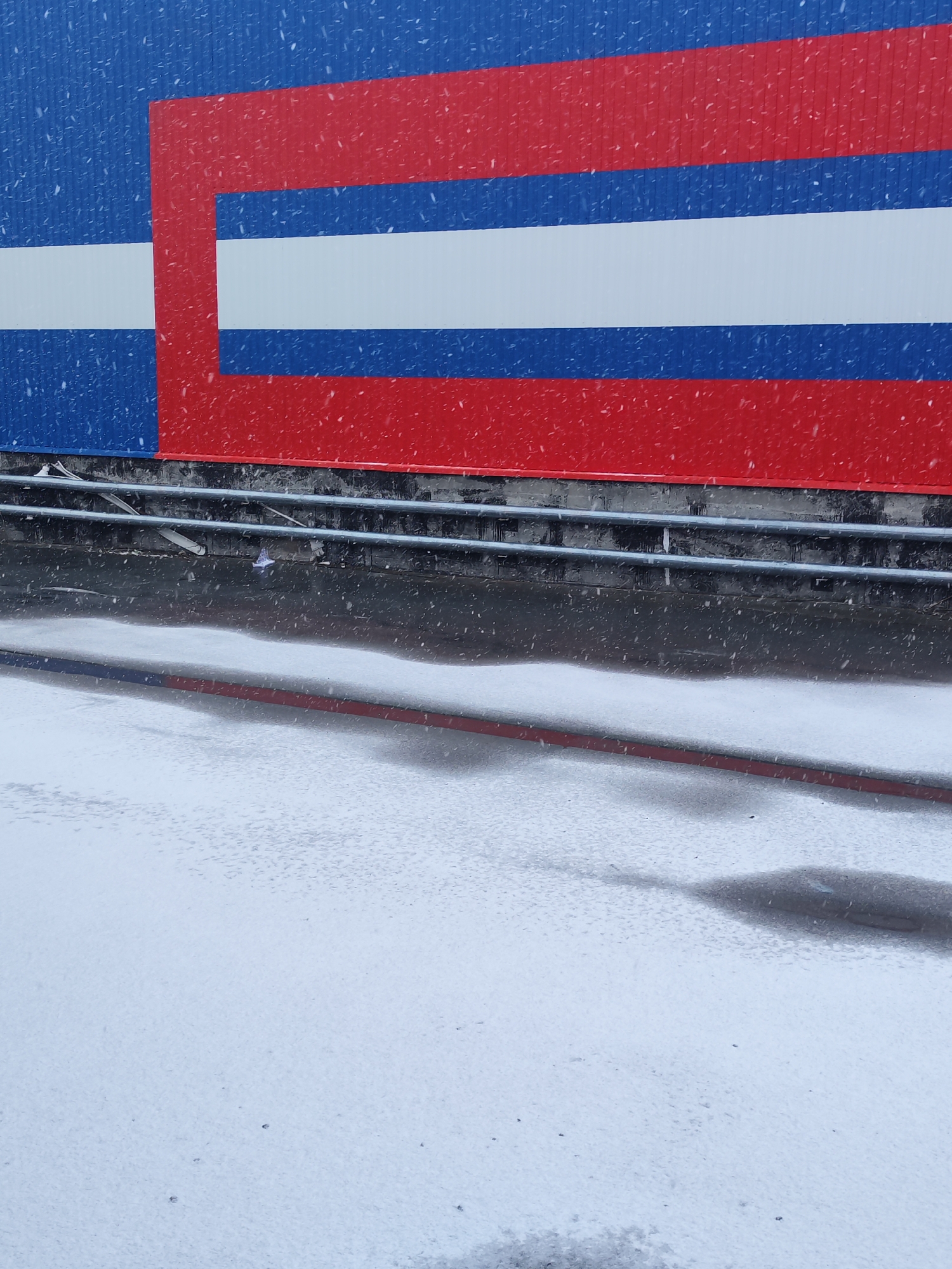 The image shows a snowy scene in Chaikovsky, Russia. A building with a blue, white, and red striped facade is visible in the background. Snow is falling and accumulating on the ground, which appears to be a paved area. There are metal railings along the base of the building. The lighting indicates an overcast day. No people are visible.