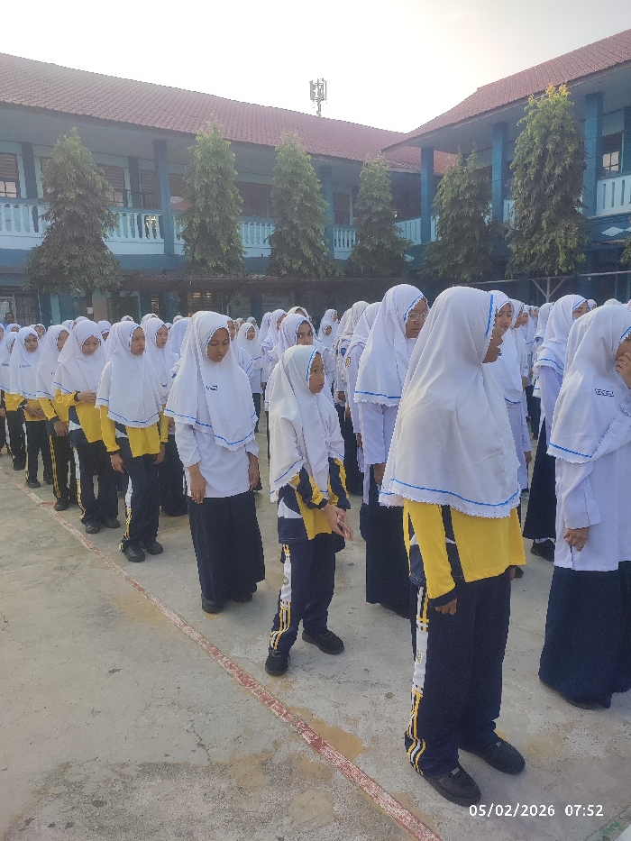 A group of young female students wearing school uniforms and white hijabs are standing in neat rows on an outdoor concrete surface. The uniforms consist of yellow tops, navy blue bottoms, and white head coverings. They appear to be lined up, possibly for a morning assembly or an organized activity. A two-story building with a red roof and blue trim is visible in the background. Trees line the back of the scene. A timestamp in the bottom right corner reads "05/02/2026 07:52".