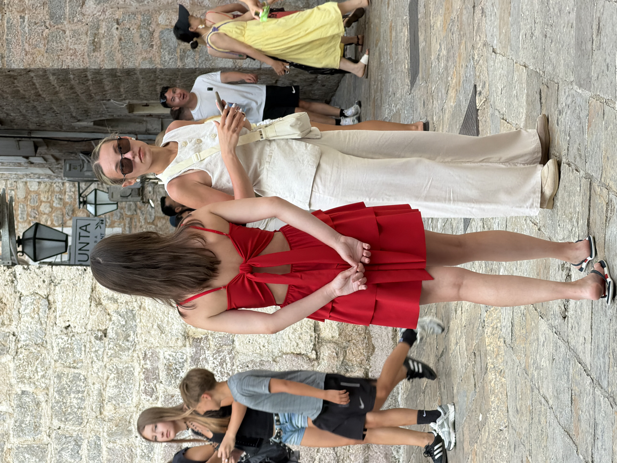 The image captures a scene on a cobblestone street in Budva, Montenegro. In the foreground, two young women are the primary subjects. The woman on the left wears a vibrant red tiered dress with a large bow detail at the back and her hands are clasped behind her. To her right, a woman in sunglasses is dressed in a white linen vest and matching wide-leg pants, holding a smartphone and a small white crossbody bag. Behind them, other individuals are visible walking through the street. To the left, a teenage girl with long brown hair and a black top stands next to a boy in a gray t-shirt and black shorts. Further back, a man in a white t-shirt and black shorts is visible. On the right side of the image, a woman in a yellow dress walks away from the camera, and another person in a dark top and hat is partially visible. The background features a stone wall with an old-fashioned lamp and signage. The overall setting appears to be a pedestrianized area with historical architecture.