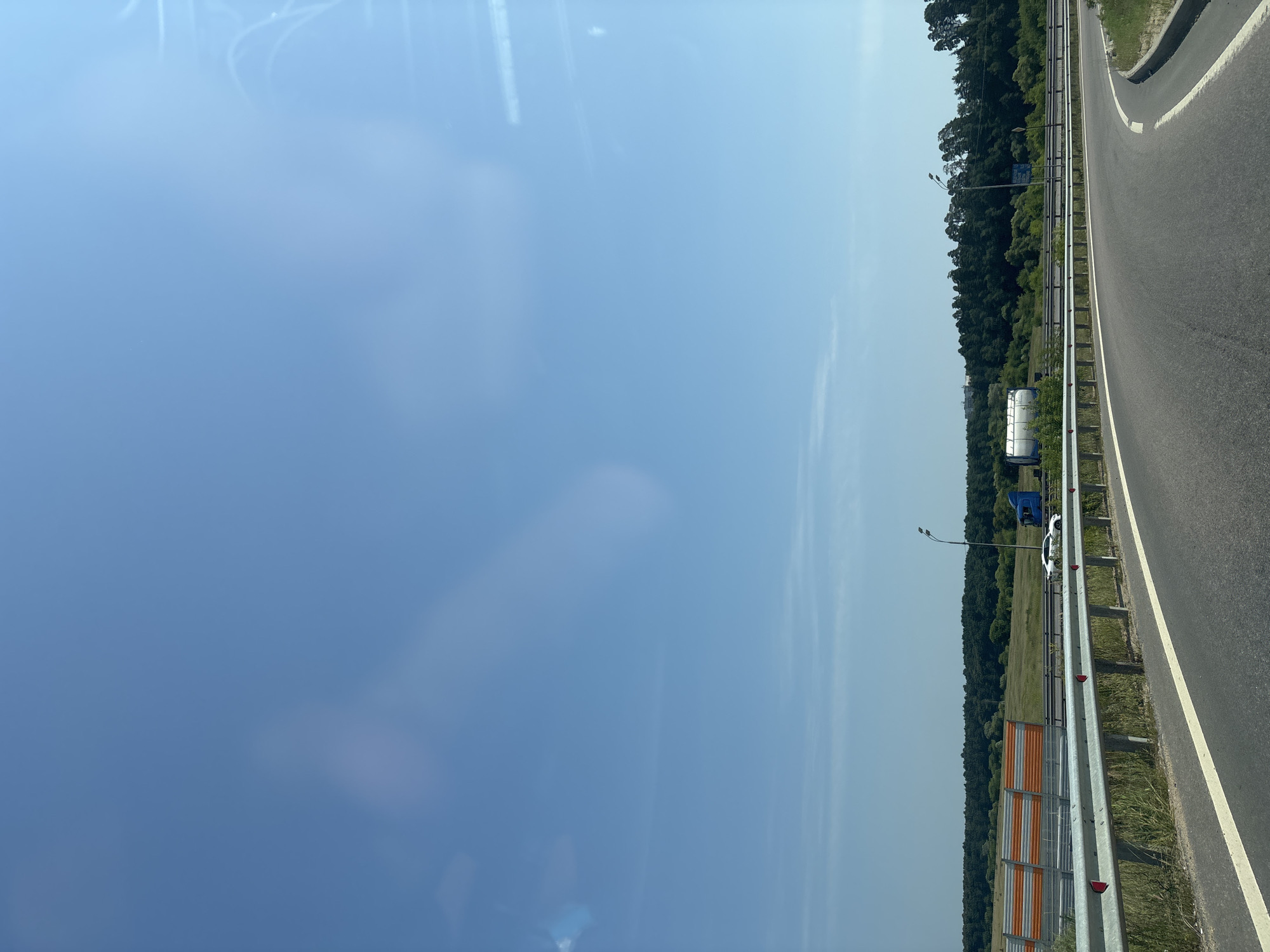 The image depicts a view from a moving vehicle on a highway, possibly in or near Zvenigorod, Russia, given the context. The sky is clear and blue, with some light cloud cover or atmospheric haze visible. The highway curves to the right, with a metal guardrail separating the lanes and a concrete barrier with orange and white noise-reducing panels on the left. Beyond the barrier, there is a grassy field and a dense forest of green trees. On the highway, a white car is visible in the distance, as is a blue truck with a tanker trailer. A single lamppost stands by the side of the road. The overall scene suggests a sunny day on a rural or semi-rural highway.