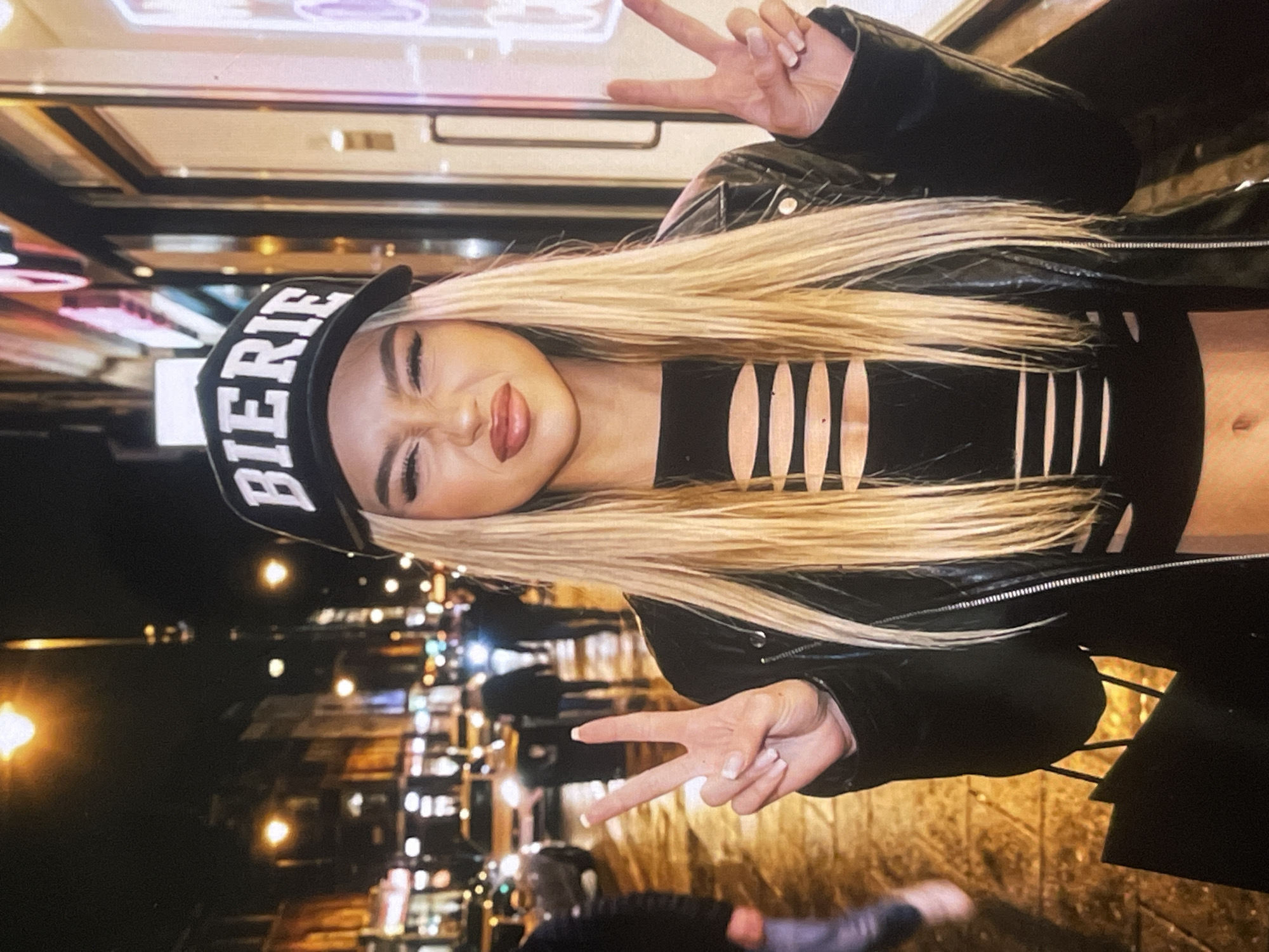 The image features a young woman posing playfully in an urban setting at night in Poltava, Ukraine. She has long, straight blonde hair and is wearing a black baseball cap with "BIERIE" printed in white capital letters on the front. Her attire consists of a black cropped top with several horizontal cut-outs across the chest, revealing her midriff, and a black leather-look jacket.
The woman is making peace signs with both hands, held up on either side of her face. Her eyes are closed or squinting, and her lips are pursed in an expressive, confident pose.
The background is dark and blurred, illuminated by numerous out-of-focus lights, indicating a city street or outdoor area after dusk. These lights appear to be from street lamps, distant buildings, and possibly vehicle headlights. Faint, blurred figures of other people can be discerned in the background, suggesting a public, lively environment. The overall impression is that of a stylish individual captured during a night out.