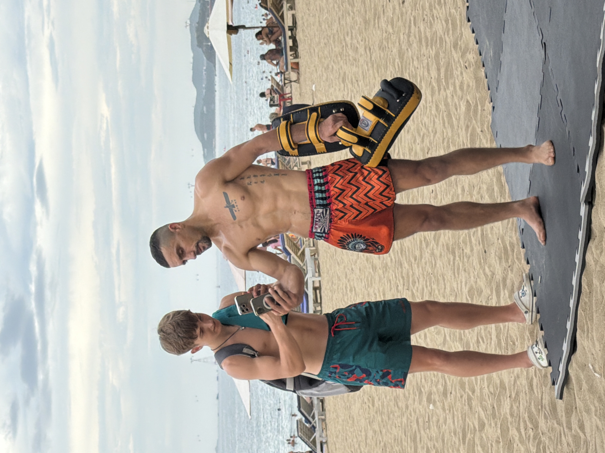 Two men are standing on a sandy beach in Nha Trang, Vietnam. The man on the left is younger, wearing dark teal swim trunks with a red and black pattern, and a black backpack. He is holding up two smartphones and looking at them. He is wearing white slip-on shoes with a floral design. The man on the right is muscular and shirtless, wearing orange and black zig-zag patterned swim trunks. He has tattoos on his chest and arm. He is holding a black and yellow Muay Thai kick pad. In the background, the ocean is visible with a cloudy sky overhead. There are beach chairs, umbrellas, and people relaxing on the beach.