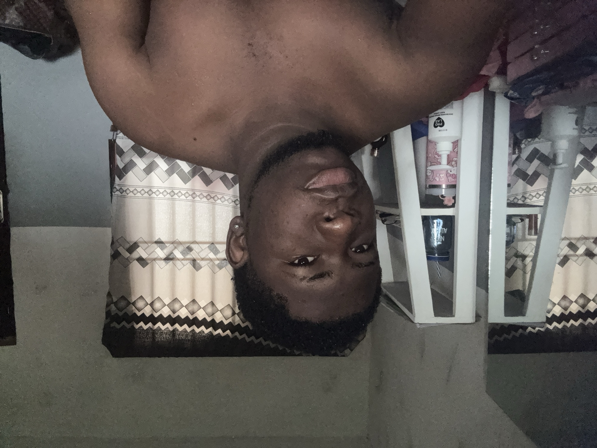 The image shows a dark-skinned man in a close-up shot, looking directly at the camera with a neutral expression. He is shirtless, revealing his torso and arms, and has a short, neatly trimmed beard and mustache. A small stud earring is visible in his left ear.
Behind the man, to his left, is a patterned curtain featuring a geometric design of diamonds and zigzag lines in white, grey, and black. To his right, a light-colored, possibly white, open shelf or cabinet is visible. On the top shelf, a white bottle with "PRO LAB" printed on it stands next to a pink and white container decorated with a "Hello Kitty" design. Below this, a dark-colored bottle, partially visible as "N AllBA" (likely part of "Natural Balance"), and some fabric items are present.
The scene takes place indoors, likely in a bedroom or living area in Ipetumodu, Nigeria, given the curtains and personal care products on the shelf. The lighting is even, suggesting indoor artificial light or diffuse daylight. The man is posing for the photograph.