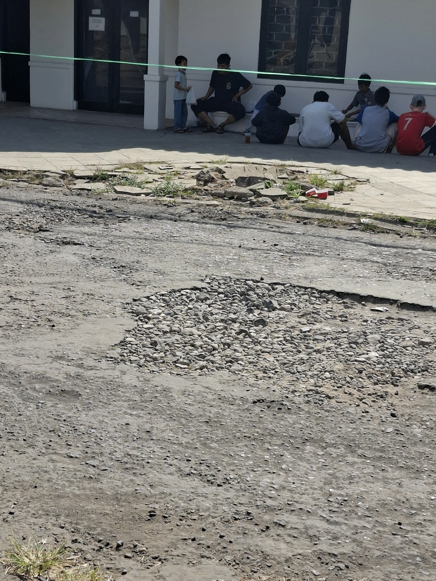 A group of young men and boys are sitting on a cracked concrete sidewalk outside a building in Karangtengah, Indonesia. A green rope is stretched across the foreground, separating the sidewalk from a heavily damaged asphalt road. The road is filled with potholes and loose gravel. Some of the individuals are wearing casual clothing, including a red t-shirt with the number 7 on the back. One young boy, standing, holds a white plastic bag. The building behind them appears to be a commercial or public establishment with large windows. The sun is shining, casting shadows.