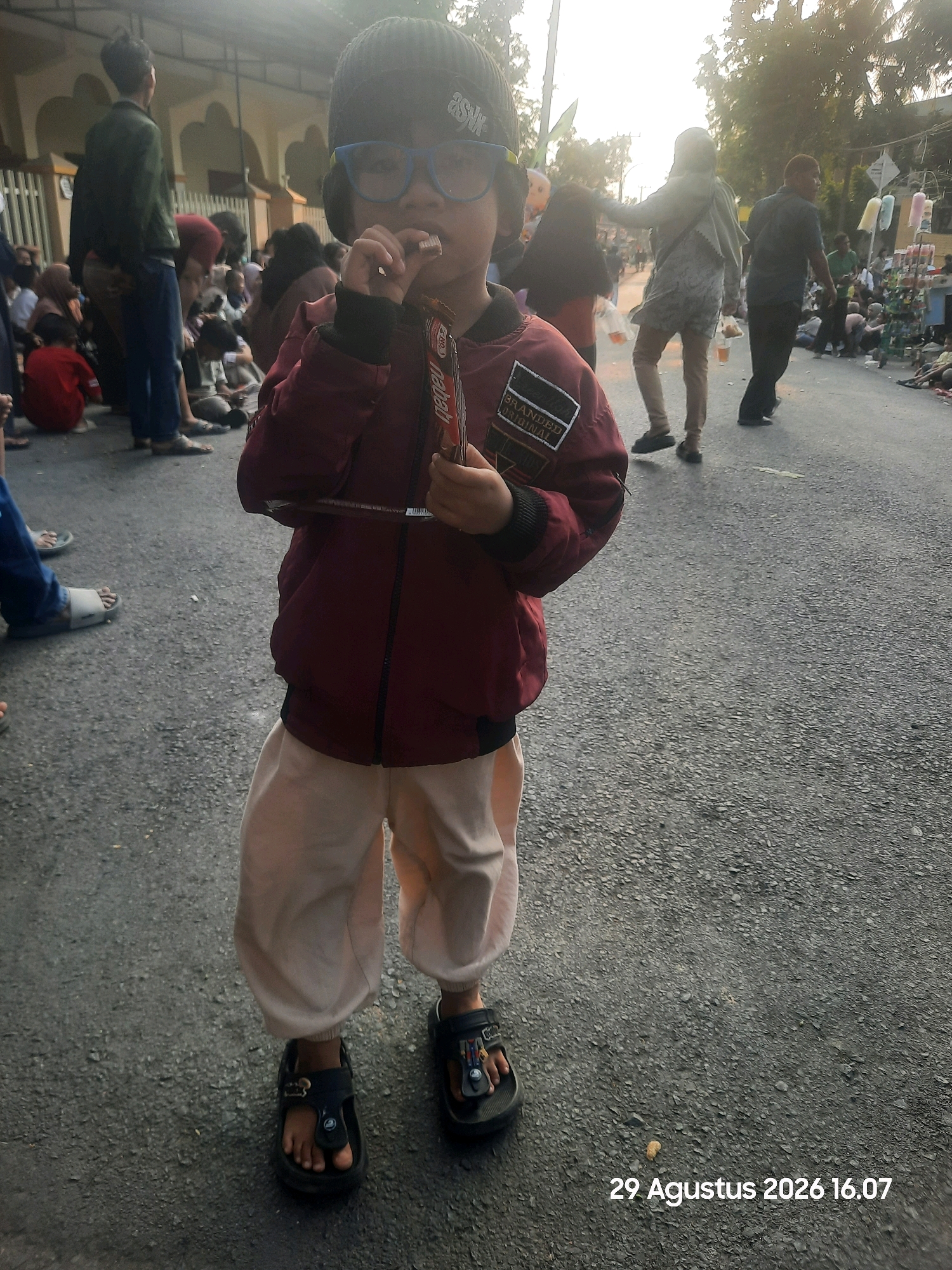 A young child wearing a dark beanie and blue-rimmed glasses is standing in the middle of an asphalt road. The child is holding a chocolate bar and eating it. They are wearing a maroon jacket with a black collar and trim, and light-colored pants that are tucked into dark sandals. The child's left foot is slightly forward, showing the details of their sandal and toes. In the background, a crowd of people are gathered, and several buildings with arches are visible. The lighting suggests it is late afternoon or early evening. The timestamp "29 Agustus 2026 16.07" is overlaid on the bottom right corner of the image, indicating the date and time it was taken. The location is identified as Patokan, Indonesia.