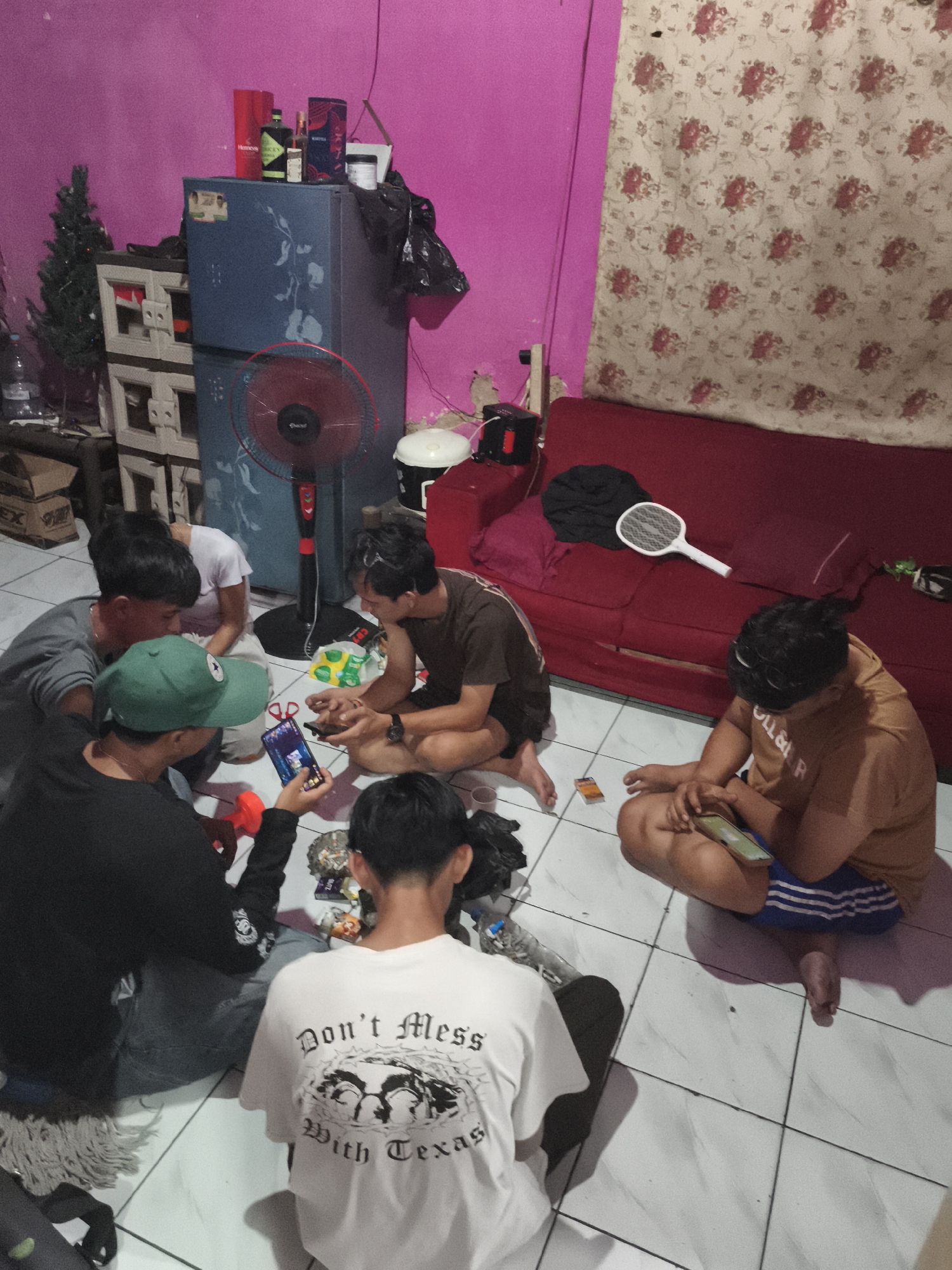In Depok, Indonesia, five young men are gathered on the floor of a room with pink walls and a patterned curtain. They appear to be engaged in a social activity, possibly playing a game on their phones or looking at something together.
The room is modestly furnished, with a blue refrigerator, a red sofa, and a floor fan visible. Several objects are scattered around the room, including bottles of alcohol, a plastic toy, and personal belongings. The overall impression is that of a casual gathering in a residential setting.