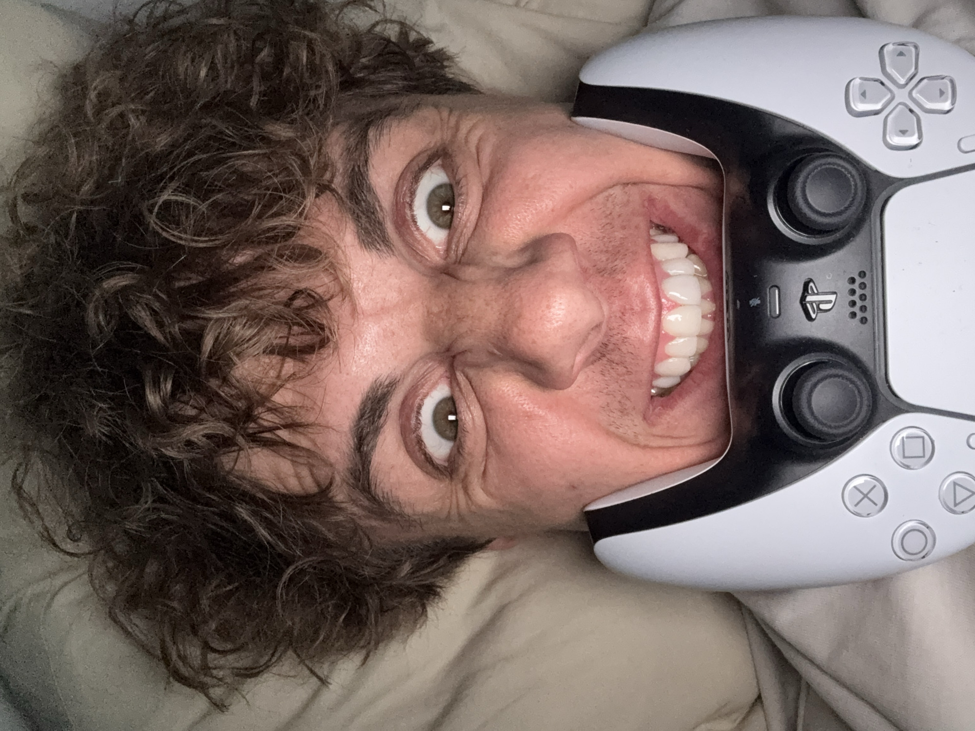 A man with curly brown hair is lying on a light-colored fabric, possibly a bedsheet. He is smiling broadly, and his face is distorted, with his nose prominently visible. He has light skin and green eyes. A white and black PlayStation 5 controller is positioned around his face, obscuring parts of his mouth and cheek. The controller's directional buttons and analog sticks are visible. The image is taken from an unusual angle, suggesting it's a selfie or taken by someone lying next to him. The scene does not provide information to locate it in Sofia, Bulgaria.