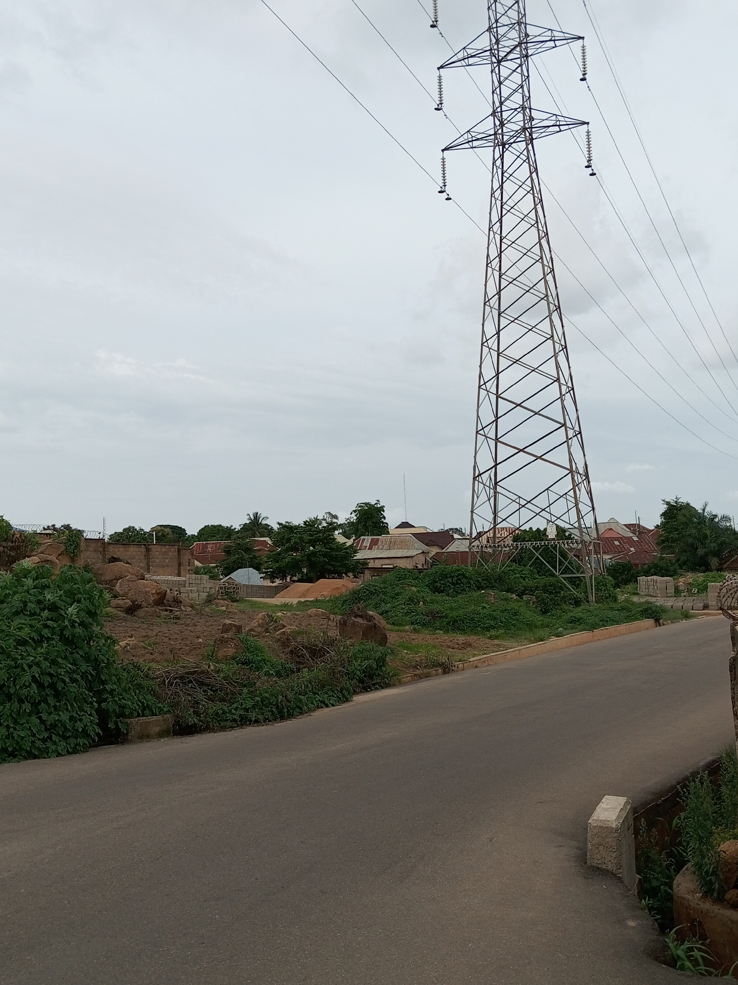 This is a medium-shot, eye-level, natural light photograph of a rural roadside in Gura Top, Nigeria. The scene is dominated by a tall, latticed metal pylon supporting high-voltage power lines, which extends upwards and out of frame. To the left of the pylon, a paved road curves away from the viewer. The roadside verge is lined with green bushes and some dirt. Beyond the road, a low brick wall encloses a plot of land. Within this plot, there are piles of sand and building materials, as well as some trees and the roofs of several buildings. The sky is overcast and a pale gray.