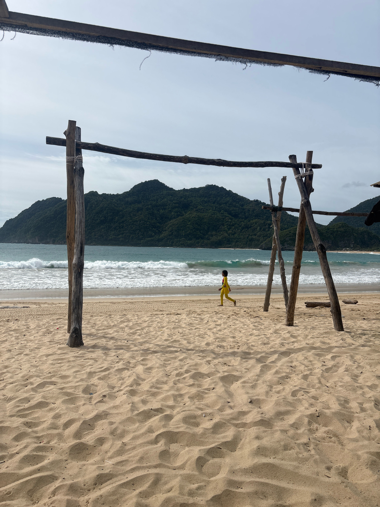 A solitary child wearing a yellow outfit walks across a sandy beach with choppy turquoise waves crashing in the background. The child is positioned to the left of the frame, facing left, with their body angled towards the ocean. In the foreground, two sets of rustic wooden posts support a rough-hewn wooden beam overhead. The sand is textured with footprints and rippled patterns. Behind the child, the ocean stretches towards a lush, green, tree-covered mountain range that dominates the horizon. The sky is overcast with a pale, diffused light. The scene appears to be a natural beach setting, possibly in a remote coastal area.