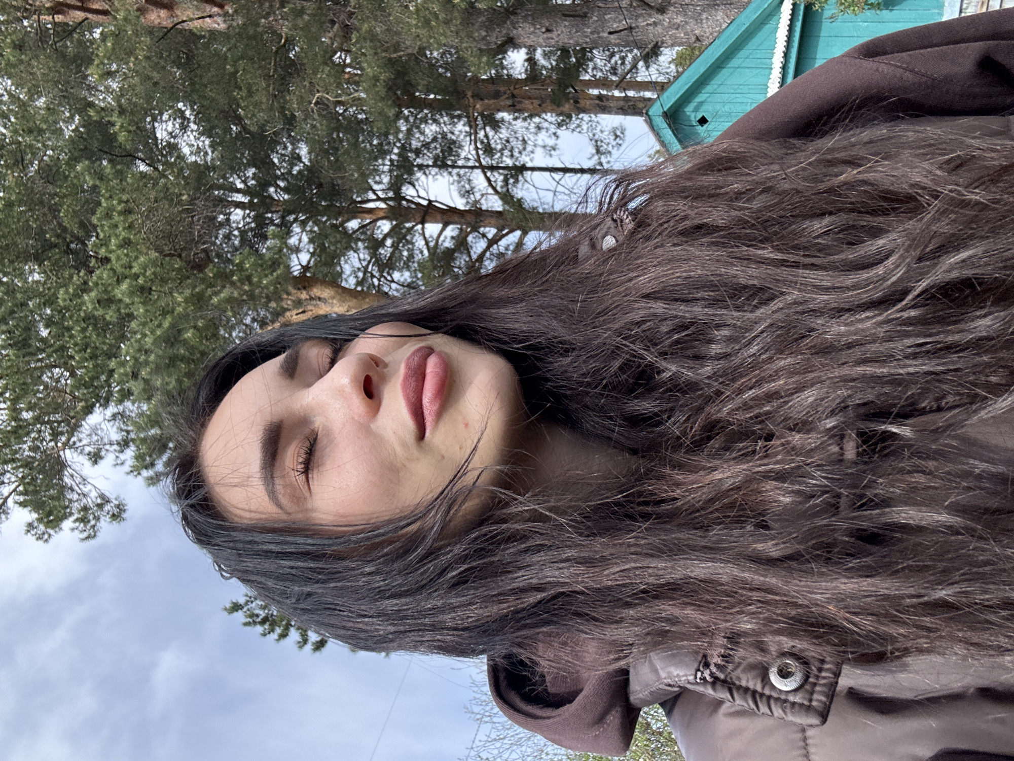 A young woman is outdoors, under trees, with a green-painted wooden building in the background. She is wearing a brown hooded jacket. Her eyes are closed. The location may be in or around the city of Chaikovsky, Russia, based on the request context.