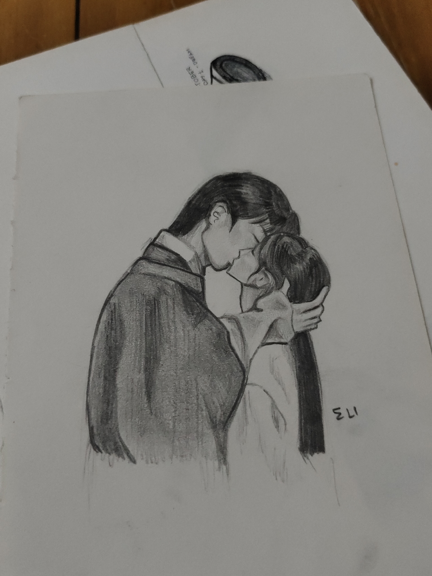 A pencil drawing on white paper depicts two stylized figures in an embrace, leaning in for a kiss. The taller figure, on the left, has dark hair and wears a dark, long-sleeved garment with a lighter collar. The shorter figure, on the right, has long dark hair and wears a light-colored, long-sleeved garment. The taller figure's left arm is around the shorter figure's mid-back, while the shorter figure's right hand rests on the back of the taller figure's head. The drawing exhibits various shades of grey, indicating pencil shading. In the upper right corner of the paper, text partially visible reads "TOBER DAY 1 - DREAM" and is oriented vertically. A signature, "ELI," is present in the lower right of the drawing. The white paper rests on a light-colored, flat surface, partially overlapping a darker, wooden surface in the upper left background. The location is an undefined indoor setting, likely a desk or table.