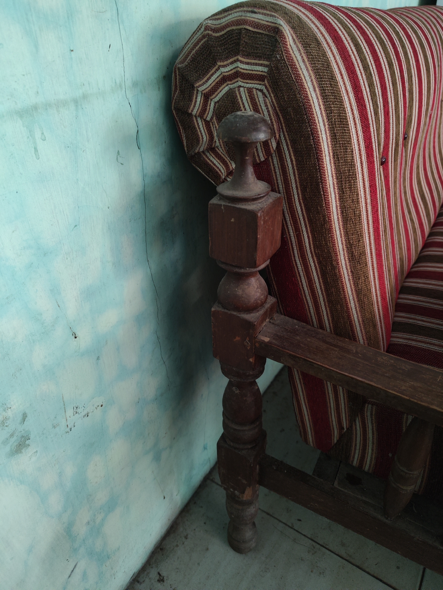 The image shows a close-up of an upholstered armchair with a striped pattern of red, brown, and off-white fabric. The armchair has a dark wooden armrest and a carved wooden finial at the top of the armrest post. The armchair is positioned next to a light blue wall that has a prominent vertical crack running down its surface. There are some water stains and minor marks on the wall. The floor beneath the chair appears to be tiled or concrete. The scene is indoors and does not contain any people or explicit indications of a specific location in Bulurejo, Indonesia, beyond the potential context provided by the user.