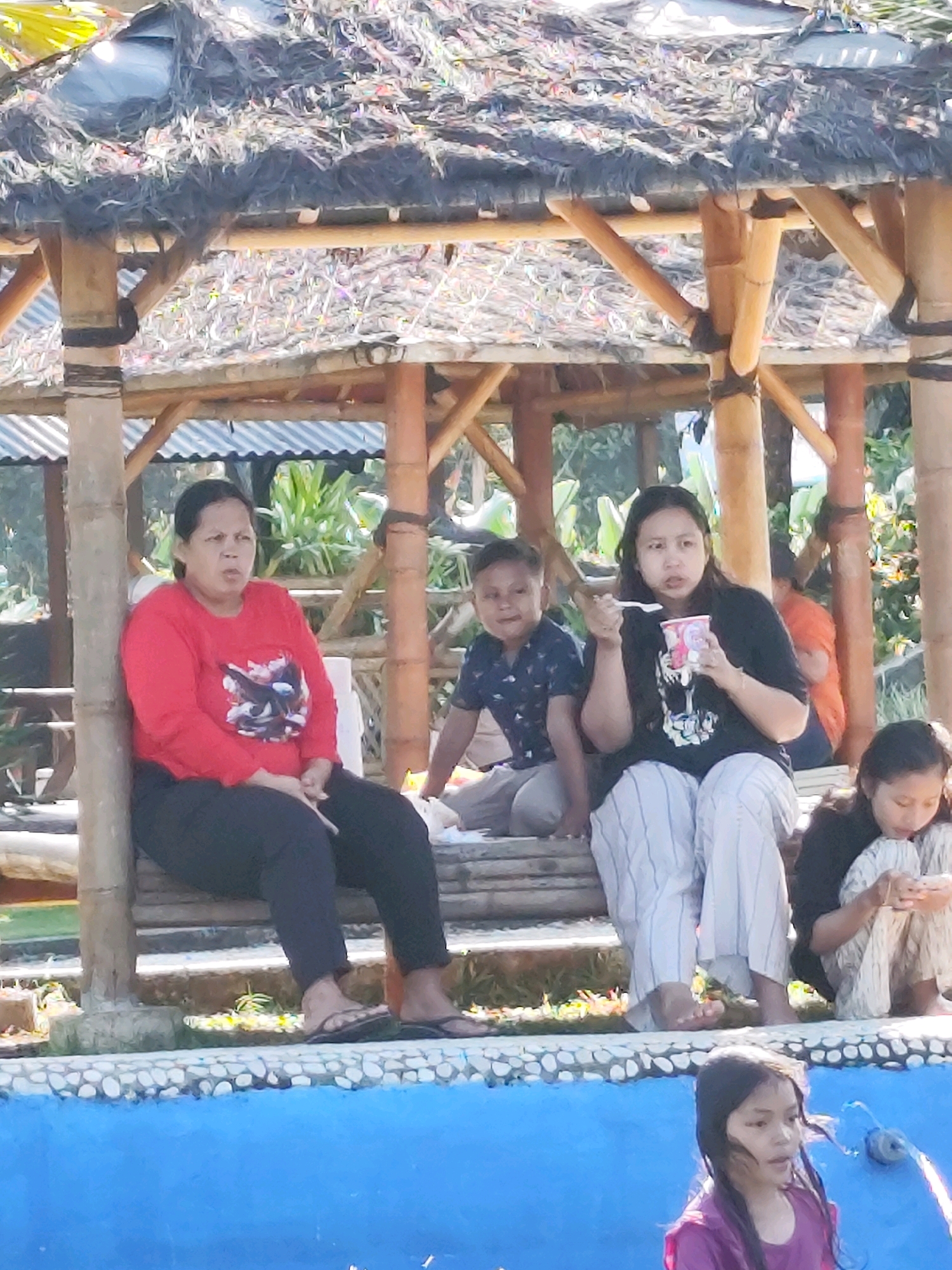In an outdoor setting with a thatched roof structure, several people are present. On a wooden bench, a woman wearing a red long-sleeved shirt and black pants sits with her legs crossed. To her right, a young boy in a dark patterned shirt and grey pants is seated. Next to him, a woman in a black t-shirt and striped pants holds a cup and a spoon. To the far right, a young girl in a patterned outfit is looking down at her phone. In the foreground, a young girl with wet hair, wearing a purple shirt, is in a blue pool. The environment appears to be a recreational area, possibly a water park or resort, with lush greenery visible in the background. The structures are made of bamboo and wood, suggesting a tropical or rustic atmosphere.