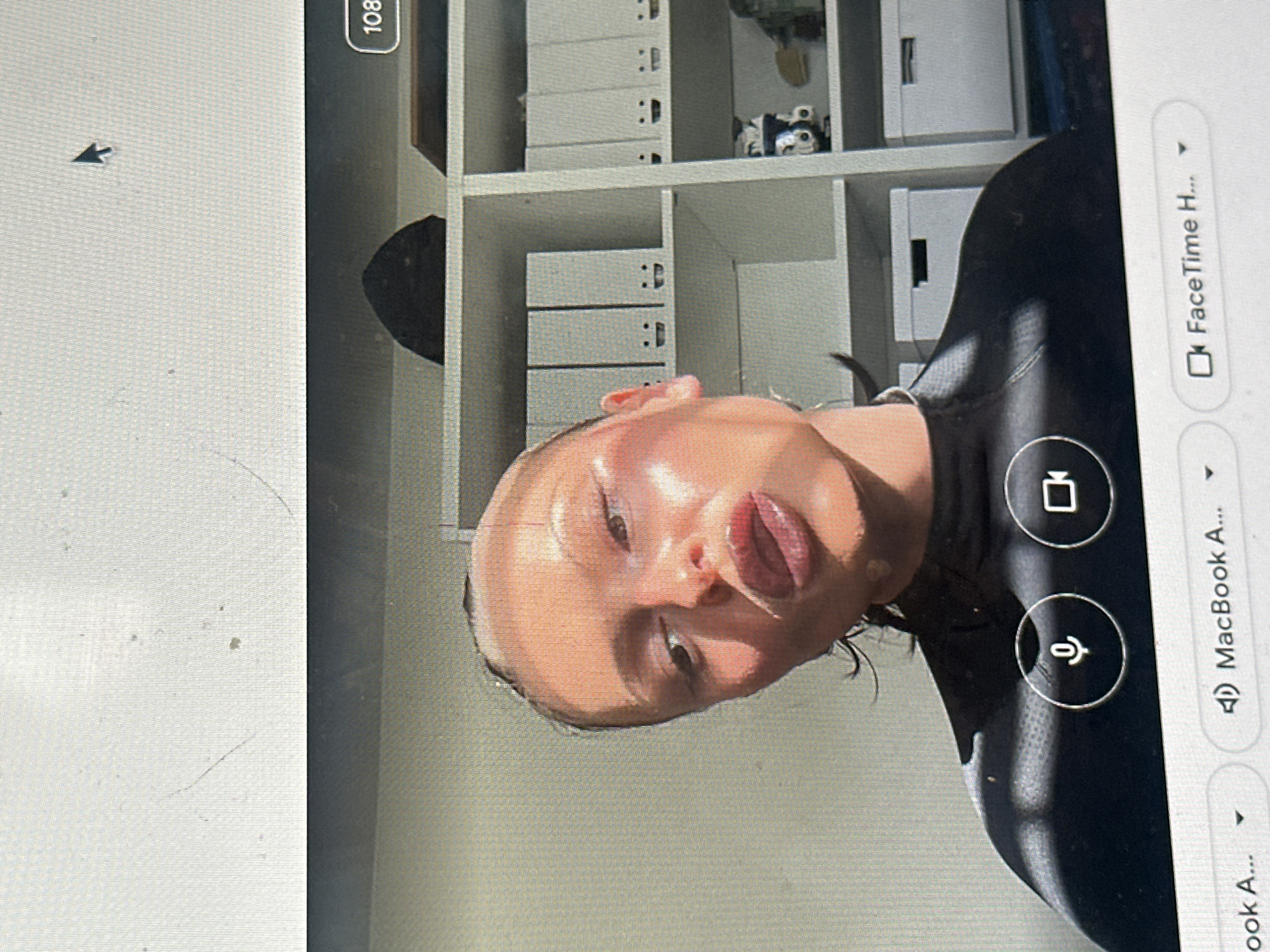 The image displays a computer screen exhibiting a video call, purportedly from a location in Wealden, United Kingdom. The screen itself shows minor surface imperfections, including visible dust specks and faint scratches, with a black mouse cursor positioned towards the upper right.
The central subject of the video call is an individual, likely female, with light skin and dark hair, visible from the shoulders up. Her face is angled slightly upwards and to the left, illuminated by strong, directional light causing prominent reflections on her forehead, cheekbones, nose, and lips. Distinct horizontal and diagonal shadow patterns are cast across her face and left shoulder, possibly from a window blind. Her lips are slightly parted. She is wearing a dark, possibly black, high-necked garment.
In the background of the video call, behind the individual, a white shelving unit is visible. The shelves hold multiple light-colored (white or grey) file boxes or binders, some marked with black smiley-face symbols on their spines. A dark, semicircular object is on the top shelf. On a lower shelf, a white storage box is present, alongside a black and white figure resembling a Stormtrooper, and other indistinct objects.
The lower portion of the computer screen displays elements of the video call interface: two white circular icons for microphone and video toggles are visible below the individual. Below these, a white bar contains text indicating "MacBook A..." with a speaker icon, and "FaceTime H..." with a video camera icon, confirming the application in use.