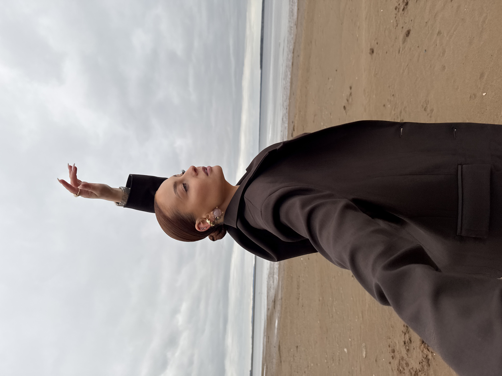 A photograph captures a person, likely a woman, striking a contemplative pose on a sandy beach under an overcast sky. The subject is positioned on the left side of the frame, from the chest up, with her head tilted back and gaze directed upwards and slightly to the left. Her reddish-brown hair is neatly styled into a bun, revealing an elaborate earring on her visible left ear, which features gold-colored elements and a translucent, possibly purple, decorative stone.
She is dressed in a dark, possibly charcoal or dark brown, long-sleeved jacket or coat. Her left arm is raised, with her hand extended upwards, showcasing a ring on her index finger and a metallic watch or bracelet on her wrist. Her expression appears serene, hinting at a moment of reflection or wonder.
The setting features a vast, light grey, cloudy sky dominating the upper portion of the image. A calm body of water stretches across the middle distance, meeting a sandy beach in the foreground. Footprints and small specks of debris are visible on the sandy surface. The overcast conditions indicate daytime, possibly on a cool or mild day. Given the context of Chaikovsky, Russia, the body of water is likely a local river or reservoir. No text is visible in the image.