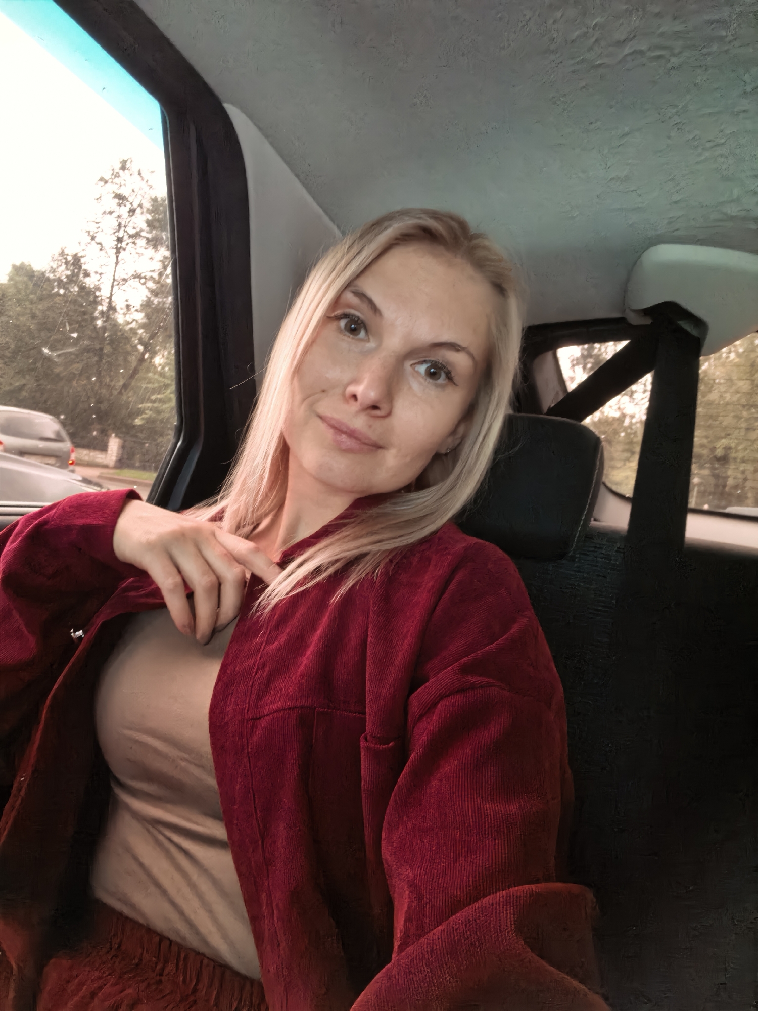 A woman with blonde hair is sitting in the back seat of a car, looking towards the camera with a slight smile. She is wearing a red corduroy jacket over a beige top. Her left hand is resting on the lapel of her jacket, partially revealing her chest. The car appears to be moving, as indicated by the blurred trees and a car in the background visible through the window. The interior of the car is visible, including a headrest and seatbelt. The lighting suggests it is daytime, possibly overcast or late afternoon. The location is not specified as Sterlitamak, Russia.