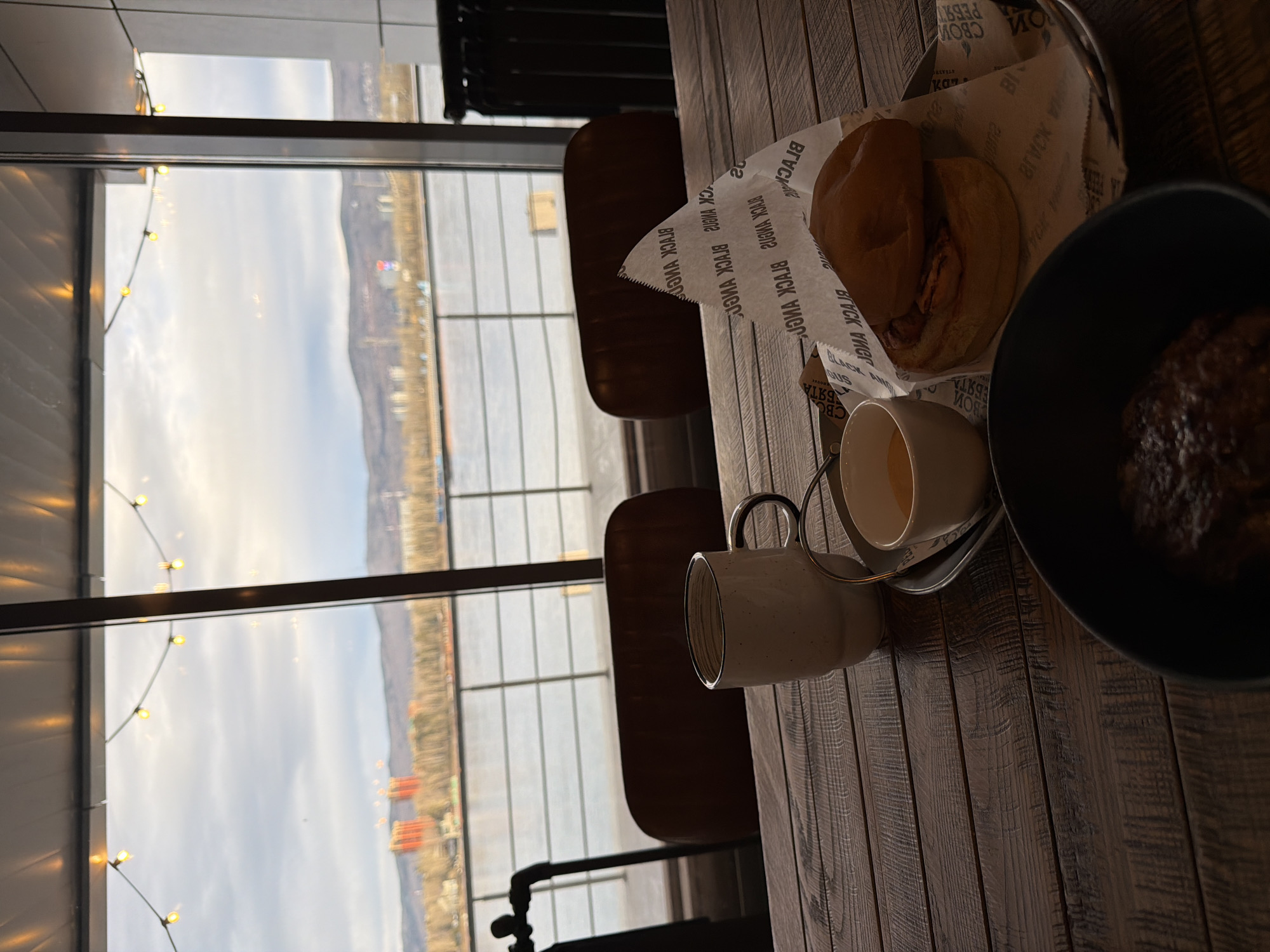 Here is a detailed description of the image:
**Overall Scene:**
The image is taken indoors, likely in a restaurant or cafe, at a table next to a large window. The table is made of dark wood planks.
**Objects:**
* **Food:** A meal is visible, consisting of two halves of a burger in a wire basket lined with paper. A dark bowl contains what appears to be a dish with gravy.
* **Drinks:** A cup of coffee or tea sits on a small saucer. Another white mug is next to it.
* **Furniture:** Two leather-upholstered chairs are partially visible.
**Window View:**
The window offers a view of a cityscape across a wide body of water, likely a river. Hills or mountains are visible on the horizon. String lights are strung across the window frame, adding a warm glow.
**Possible Location:**
Based on the river, skyline, and hills in the background, it is highly probable that the location is Krasnoyarsk, Russia. The Yenisei River flows through Krasnoyarsk, and the city has a distinctive mountainous backdrop.
**Overall Impression:**
The scene conveys a sense of casual dining with a scenic view. The lighting suggests either early morning or late afternoon. The focus is primarily on the food and the ambiance provided by the window view.