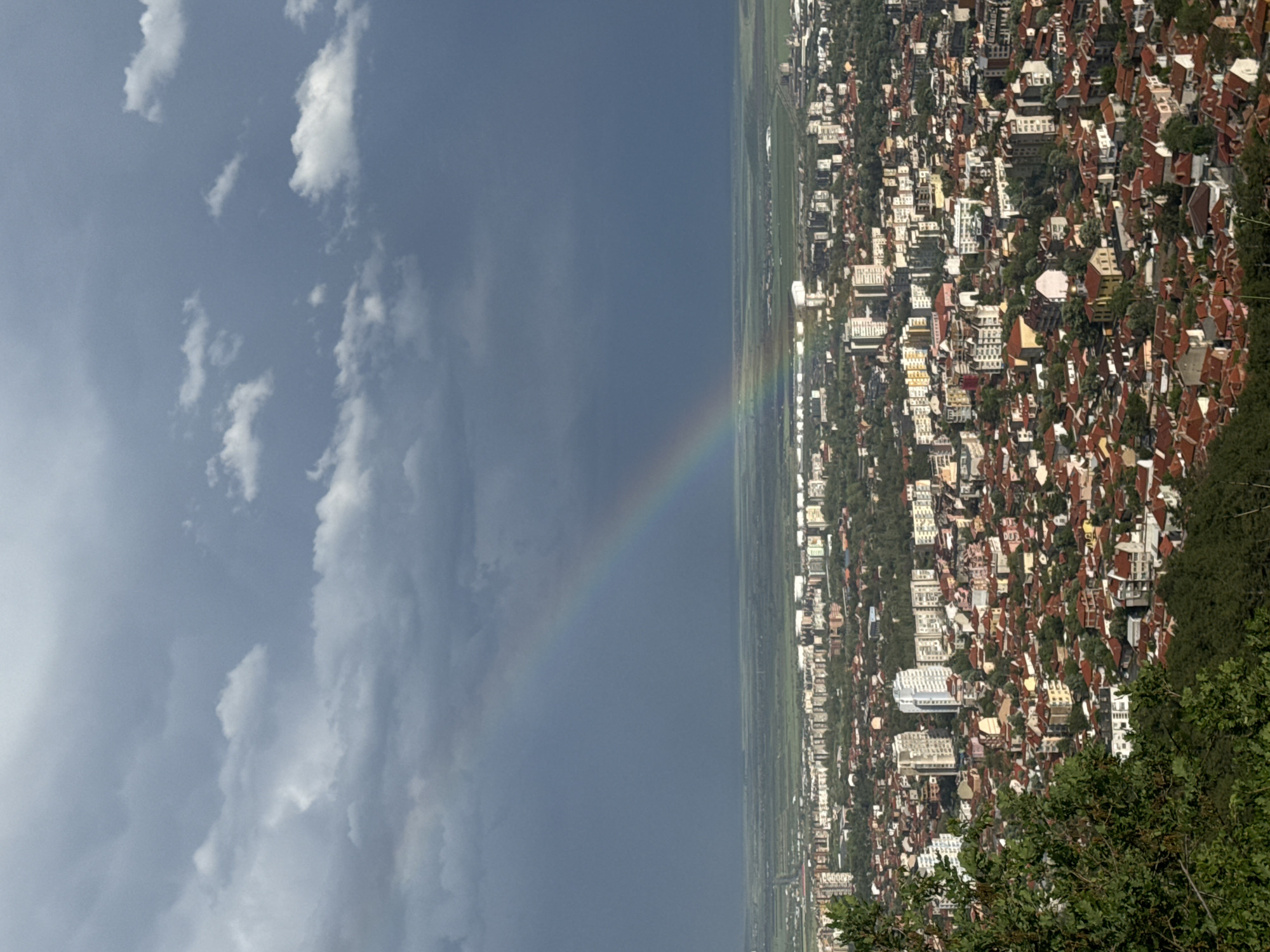 The image presents an aerial view of a city landscape under a cloudy sky, featuring a faint rainbow arcing across the scene. The right side of the frame showcases a dense urban area with numerous buildings, many characterized by red-tiled roofs. Green trees are interspersed among the structures. To the left, the sky is overcast with various cloud formations. A faint rainbow spans from the urban area into the darker sky, adding a touch of color to the scene. The location appears to be in an urban or suburban area, with a mix of residential and commercial buildings. The overall atmosphere suggests a recent rain event that gave rise to the rainbow.