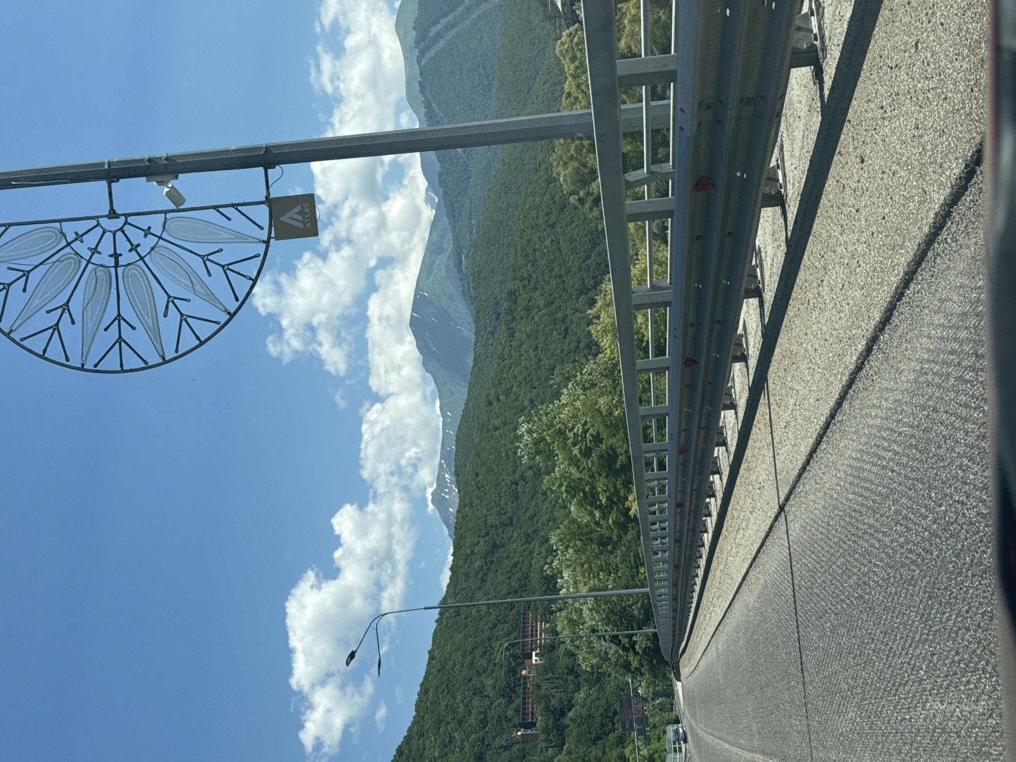 The image shows a view from a vehicle driving on a bridge in Krasnaya Polyana, Russia. The bridge has a metal guardrail with supporting posts. On the right side of the bridge, a tall metal pole holds a decorative, semi-circular metal ornament resembling a stylized flower or sun, along with a security camera and a small square sign with the word "ATLAS" printed on it. In the background, lush green forested mountains rise under a bright blue sky with scattered white clouds. Several snow-capped peaks are visible in the distance. On the left side of the bridge, a road curves away, leading towards a cluster of buildings nestled on the mountainside. Two streetlights are also visible on the left side of the bridge. The road surface is asphalt, with visible expansion joints and a textured aggregate shoulder.