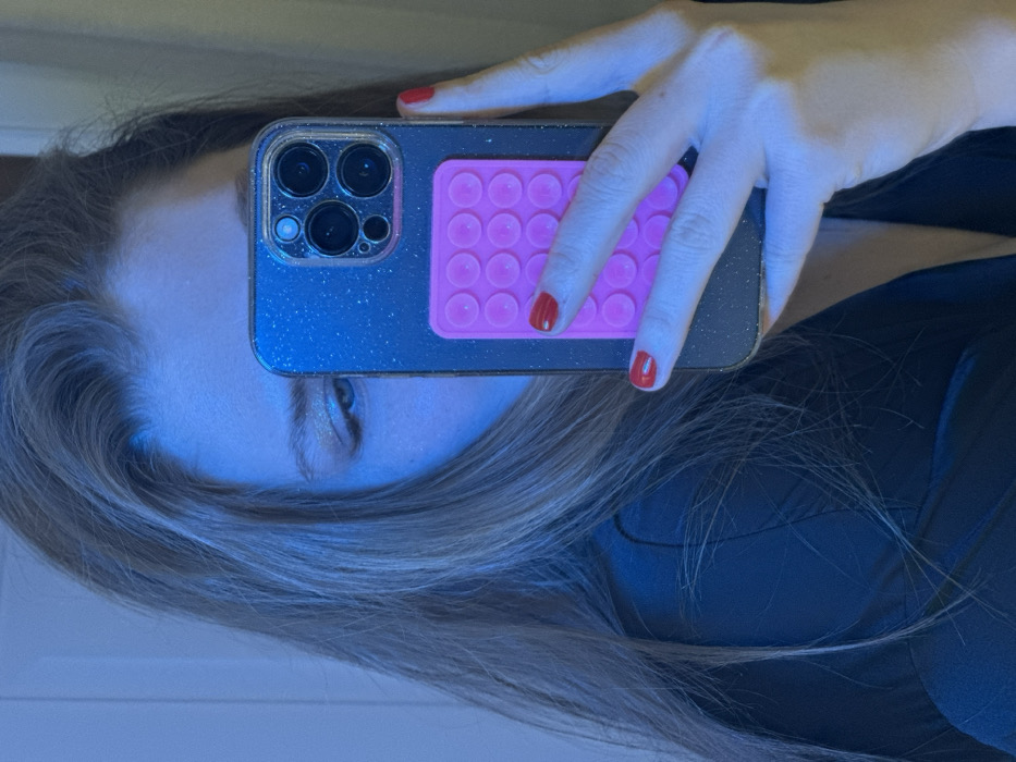A person is holding a phone in front of their face, obscuring most features. The phone has a sparkly blue case and a pink, rectangular pop-it attached to the back. Red nail polish is visible on the fingers holding the phone. Only one eye is visible, appearing light in color. The person has light brown or blonde hair. They are wearing a dark-colored top. The image is rotated to the side.
