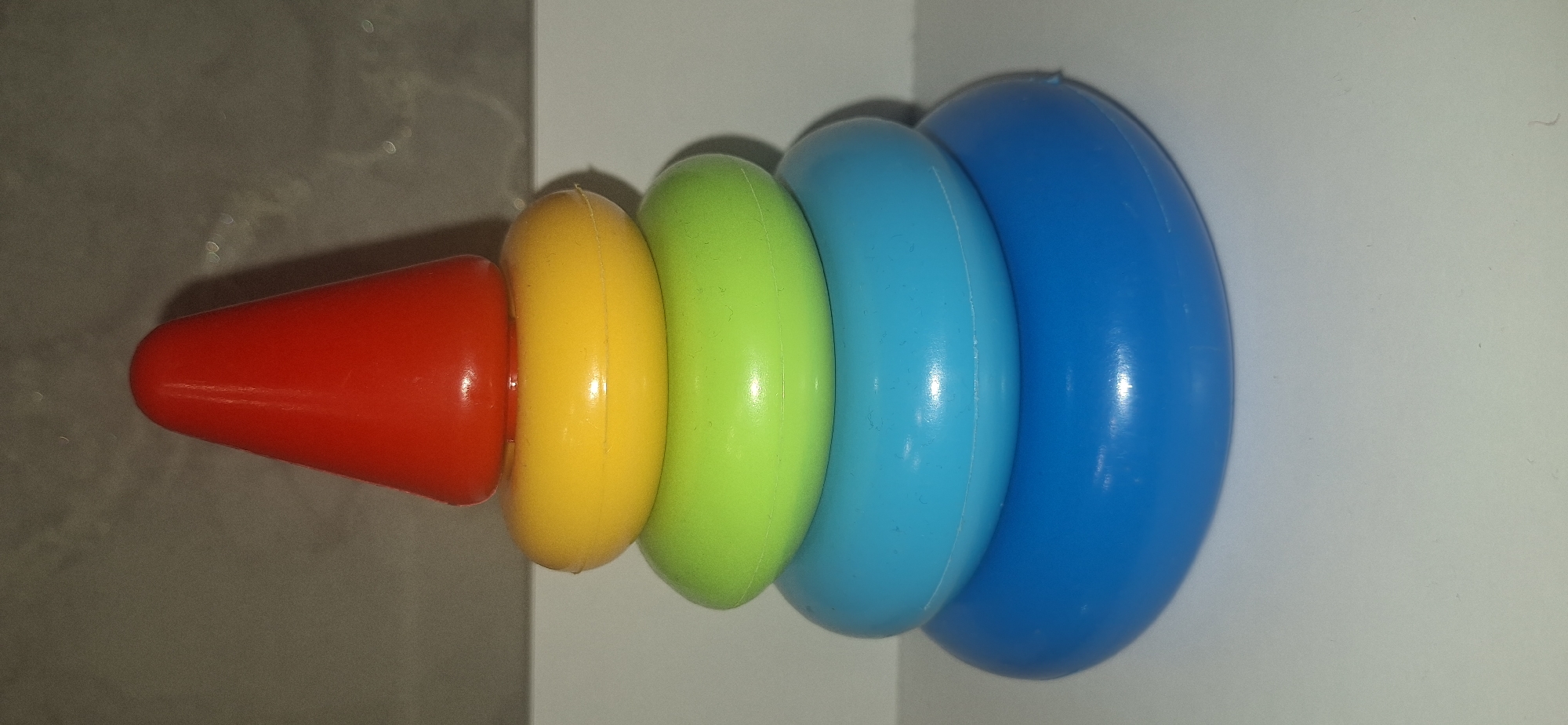 A colorful stacking toy is displayed on a white surface against a neutral gray background. The toy consists of five parts. At the base is the largest, bright blue ring. Stacked on top of that is a slightly smaller, medium blue ring, followed by a green ring. The next ring is yellow, and the topmost piece is a red cone. The lighting casts shadows from the toy onto the white surface. There is no indication of the city of Bila Tserkva, Ukraine, or any specific location. The image focuses solely on the toy.