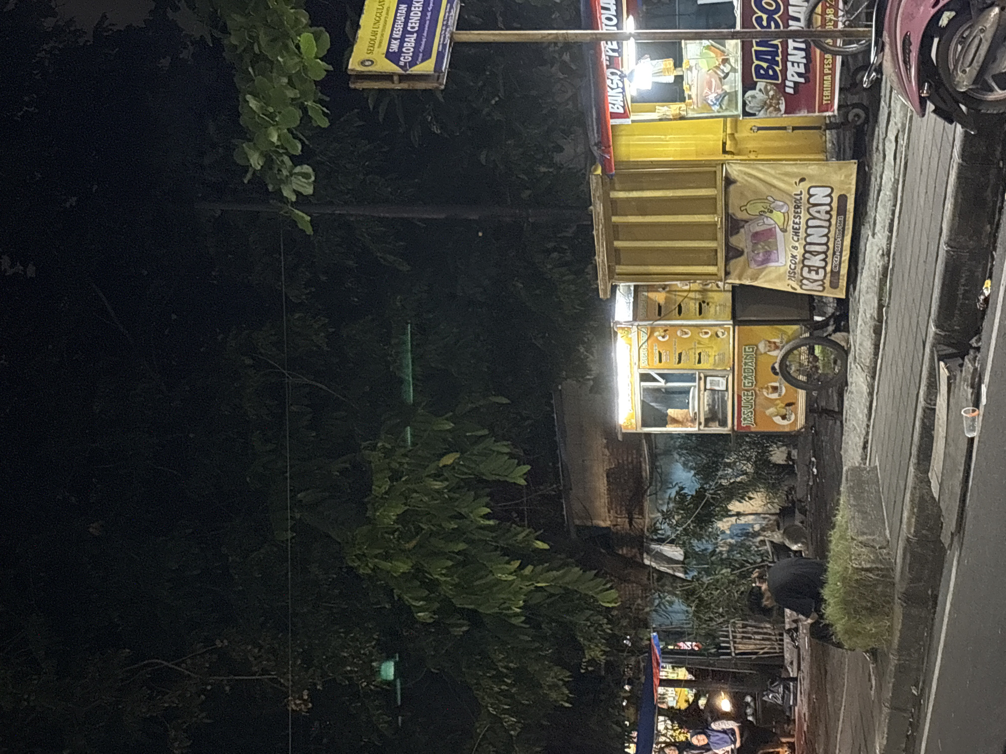 At night in North Jakarta, Indonesia, a row of street food vendors line the edge of the pavement. Each vendor has a cart with signage, including a yellow cart advertising "JASUKE GADANG," a cart with "PISCOK & CHEESEROLL KEKINIAN," and a cart with "BAKSO PE TOLA." A sign for "SMK KESEHATAN GLOBAL CENDEKI" is visible. A person is seated near the carts, and other people are gathered in the background. A parked motorbike is visible at the edge of the frame.