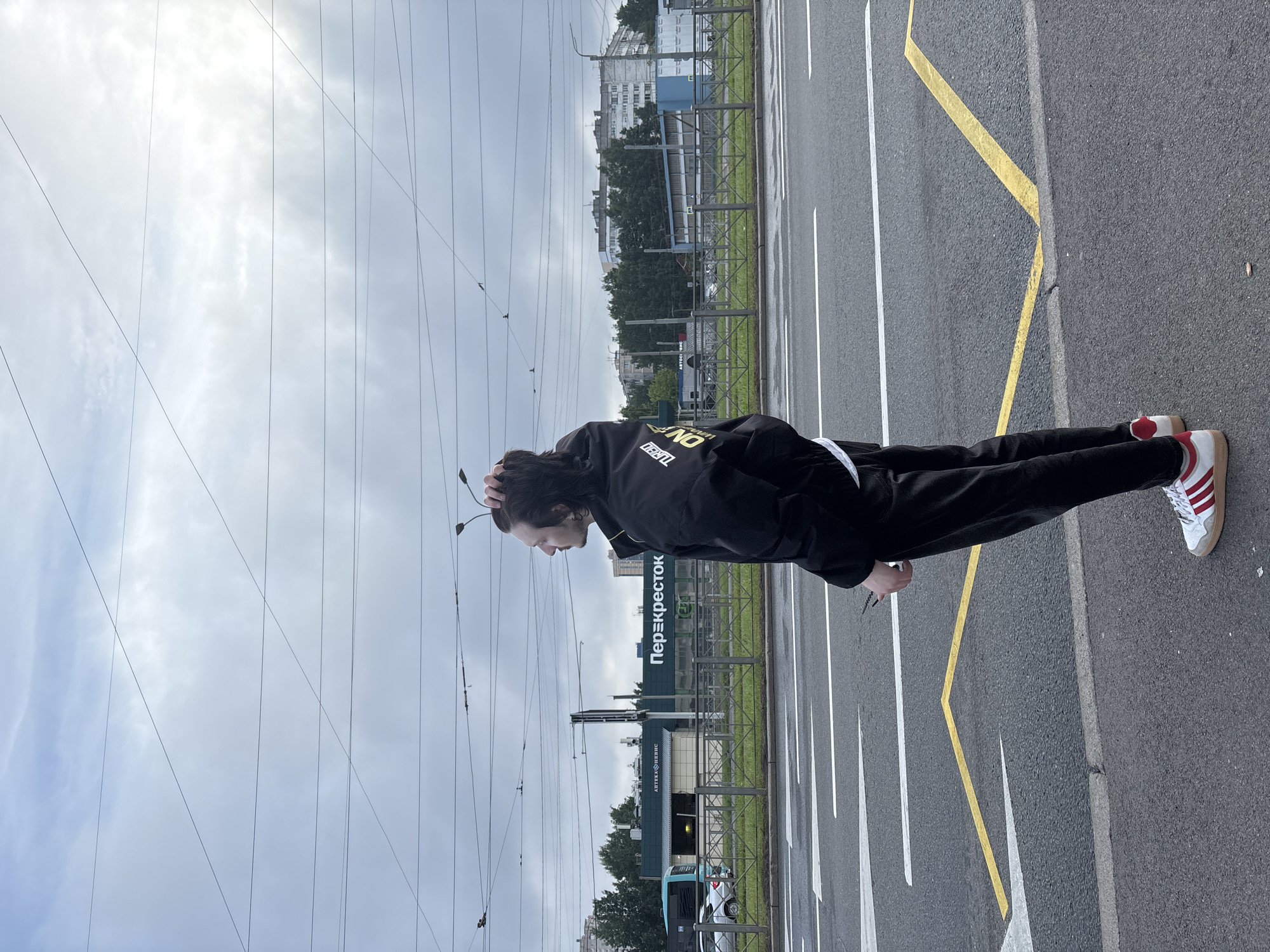 A young man stands on a street in Saint Petersburg, Russia, looking to the side with his hand on his head. He is wearing a black jacket and black pants, with white and red sneakers. In the background, there is a street with yellow and white markings, overhead wires, buildings, and trees. A supermarket with the sign "Перекрёсток" is visible on the left. The sky is cloudy.