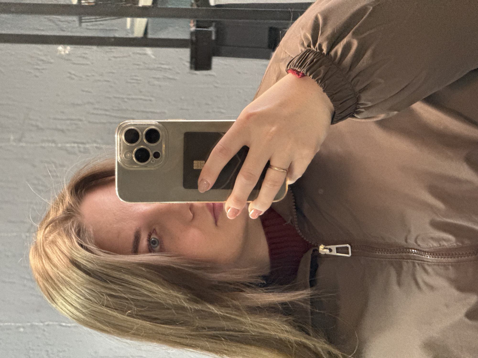 A young adult female is depicted in a mirror selfie, taken indoors in Chaikovsky, Russia. Her face is partially obscured by a light-colored smartphone held in her right hand, featuring a visible triple-lens camera array and a clear case. A dark card is partially visible behind the phone in her grip. The subject has long, light brown hair, a visible blue eye, and manicured nails with light pink polish. She wears a dark red turtleneck sweater beneath a brown zippered jacket with elasticized cuffs. A red string bracelet is present on her right wrist, and a plain gold-colored ring is on her right ring finger. The background consists of a textured, light gray wall, with reflections in the mirror showing dark vertical structures and lighter surfaces to the right, indicative of an interior environment.