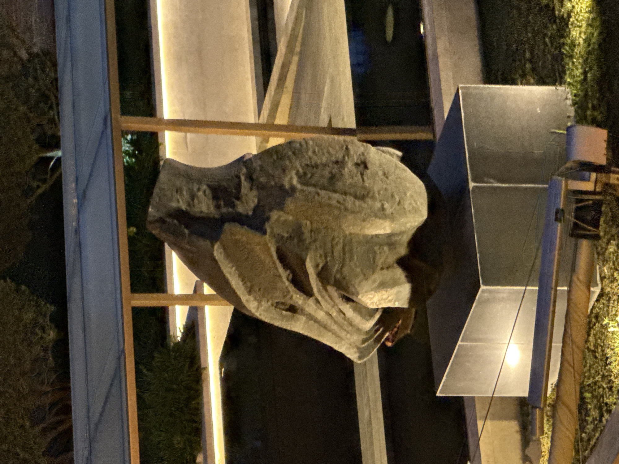 The image depicts an outdoor sculpture in Baku, Azerbaijan. The sculpture, composed of rough, light-colored stone, is mounted on a cubic pedestal. It is illuminated by artificial lighting, casting shadows and highlighting its textured surface. Behind the sculpture, a modern architectural structure with a sloped roof and supporting beams is visible, further enhanced by warm-toned lights. The background features dark greenery and trees, suggesting a nighttime setting within a landscaped area. The overall scene suggests an artistic installation in a contemporary urban environment.