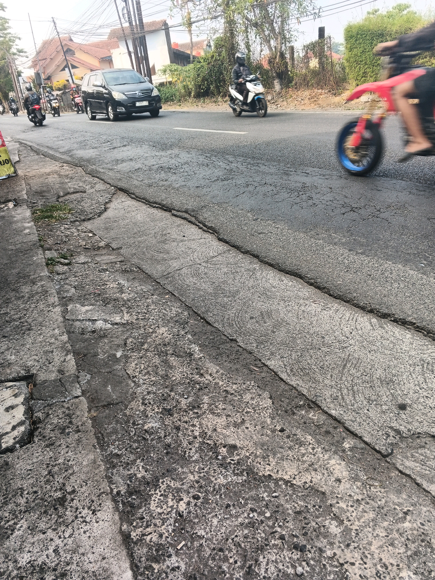 The image shows a street scene in Ungaran, Indonesia. A black SUV is driving on the road, followed by several motorcycles. A person on a red motorcycle with a blue wheel is in the foreground, partially blurred due to motion. The road is asphalt with cracks and patches, and there is a concrete curb on the left side of the image. Buildings with terracotta roofs and utility poles with electrical wires are visible in the background. The weather appears to be clear and sunny.