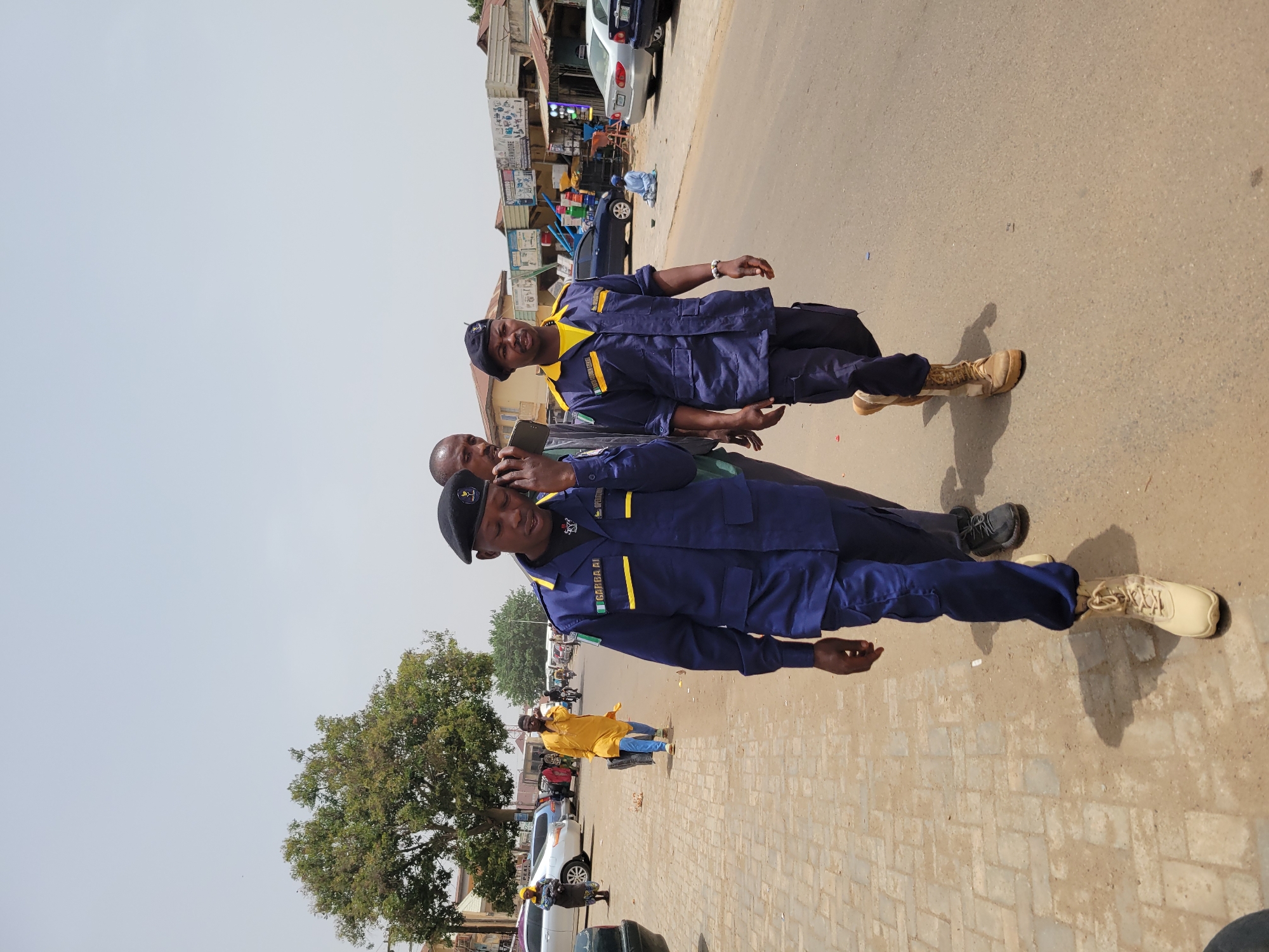 The image shows two men in dark blue uniforms with yellow trim, possibly security personnel or law enforcement officers, standing on a paved street in Jos, Nigeria. One man wears a black beret and has a name tag that reads "GARBA AI". He appears to be holding a phone to his ear. The other man stands behind him, also in uniform. They are located on a street with buildings and shops in the background. Cars are parked along the street. A tree is visible to the left, and other people are visible in the distance. The environment appears to be a busy commercial area.